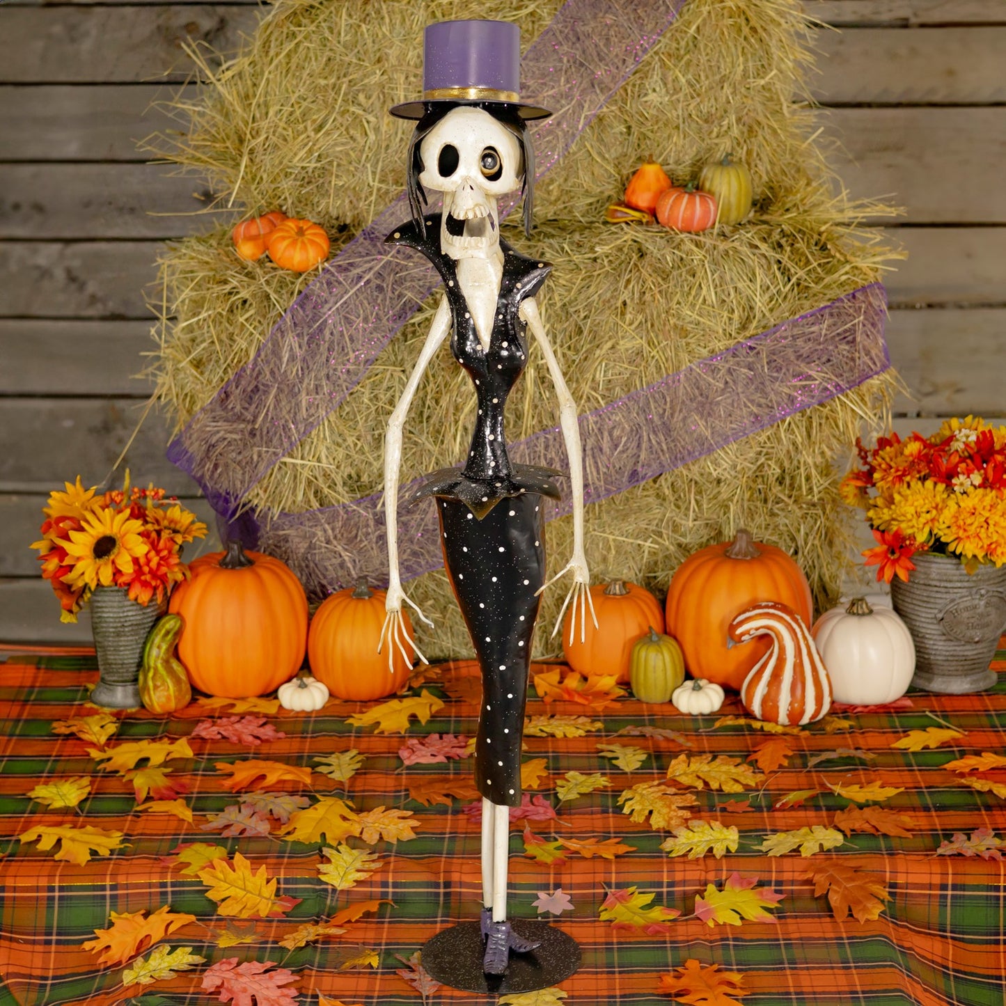One Halloween skeleton figurine dressed in black outfit, standing on a plaid autumn tablecloth with pumpkins, flowers, and a hay bale backdrop.