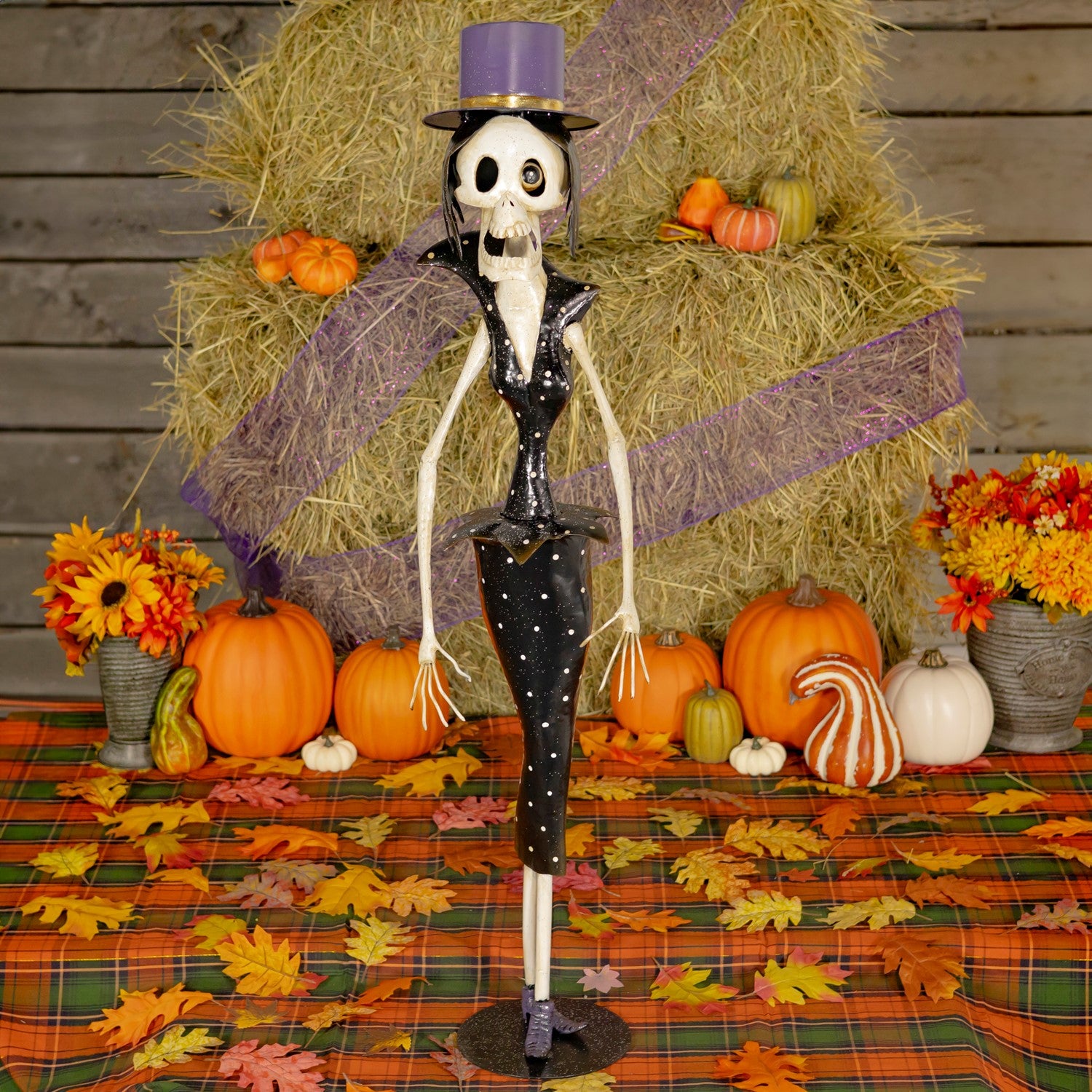 One Halloween skeleton figurine dressed in black outfit, standing on a plaid autumn tablecloth with pumpkins, flowers, and a hay bale backdrop.