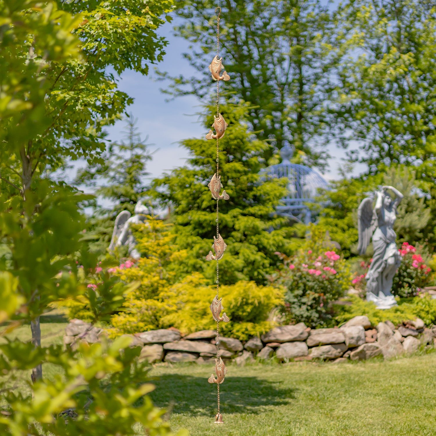 A garden scene with a hanging string of small dried flower petals or seed pods suspended vertically in the center. The background shows green trees, grass, a stone wall, and gray angel statues under a blue sky.
