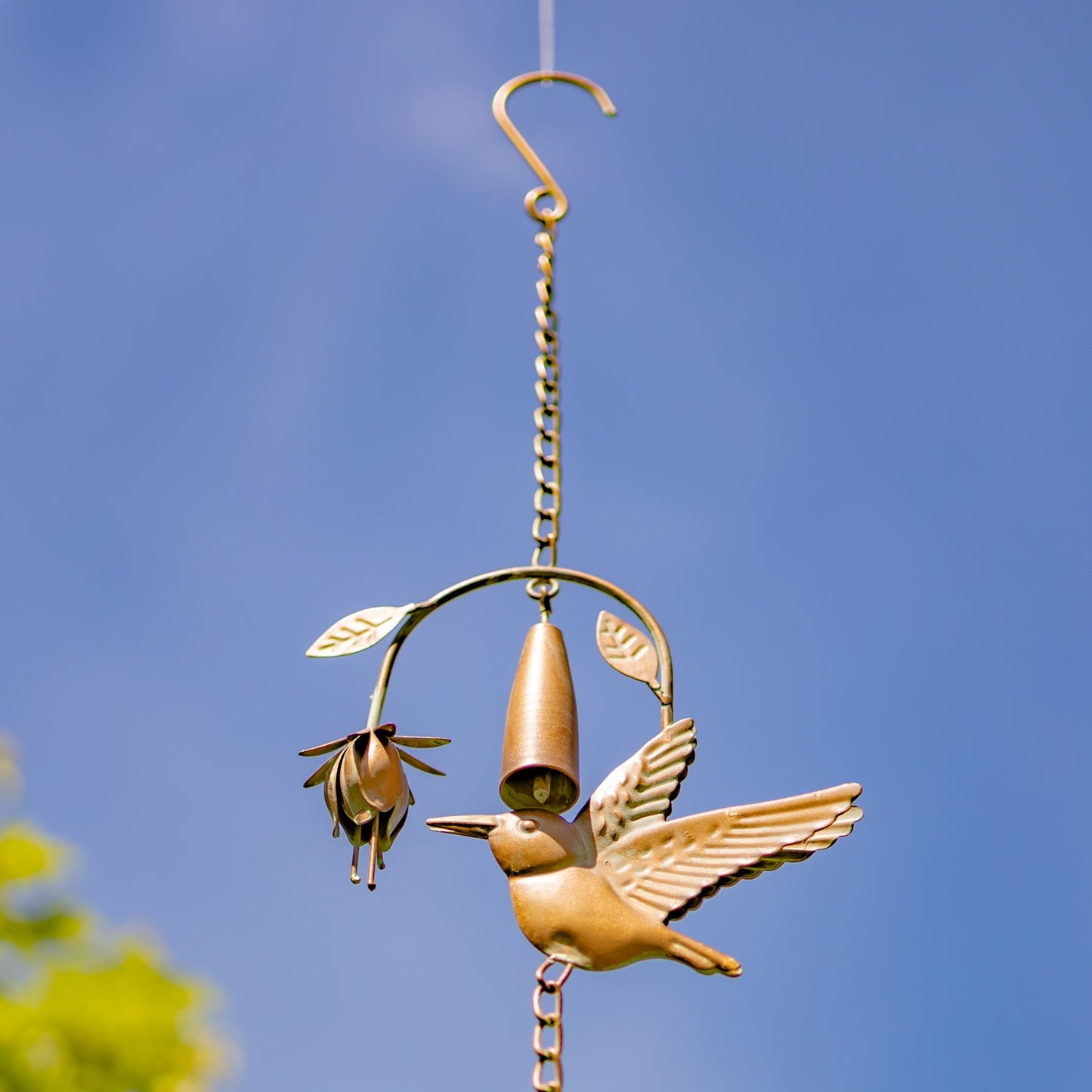 A bronze bird feeder hanging outdoors against a clear blue sky, featuring a bird figure, feeder cup, and small flower with leaf details.