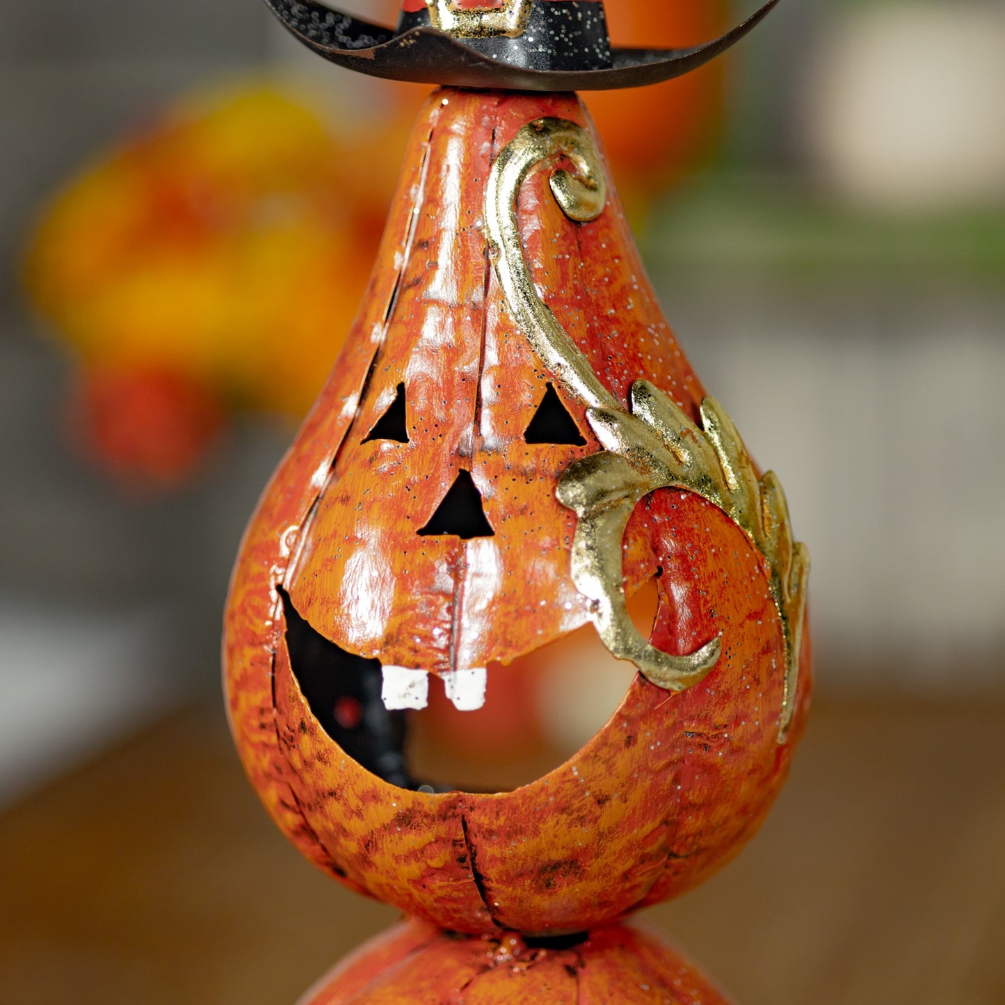 A close-up of a decorative gourd-shaped jack-o'-lantern with a carved face, a black witch hat at the top, and gold-toned scroll accents on a warm orange-brown background.
