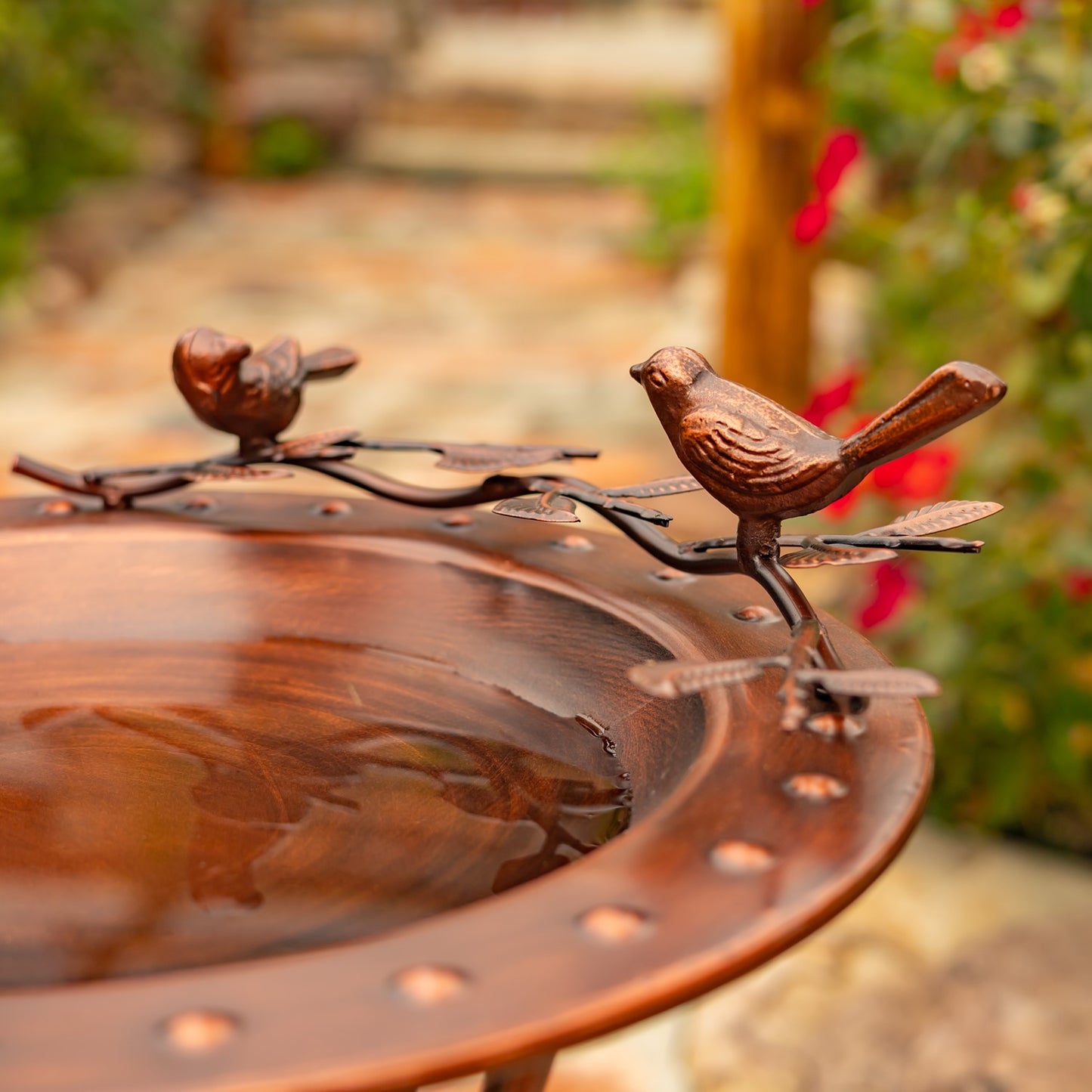 A copper bird bath with decorative metal birds perched on the rim, shown outdoors with a blurred garden background.