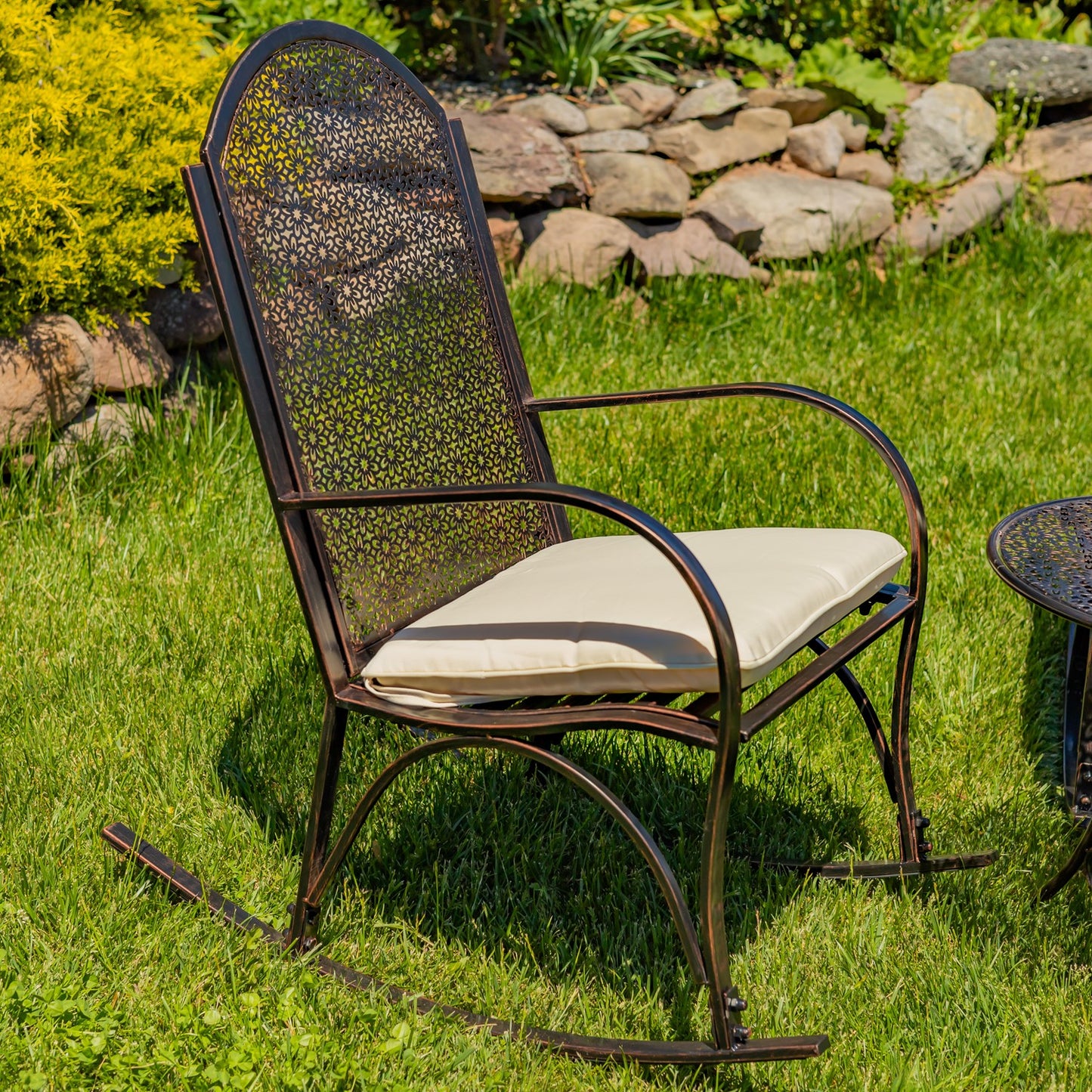 A dark brown metal rocking chair with a woven cane backrest and a cream cushion, set on a green grassy lawn with a stone garden bed in the background.