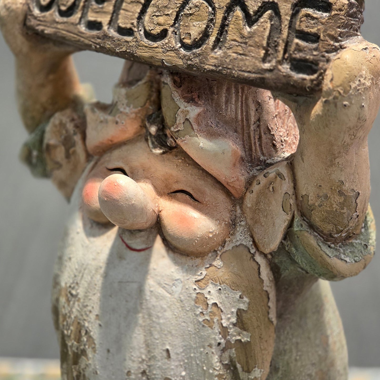 Close-up of a weathered gnome garden statue holding a carved wooden sign that reads "WELCOME". The sculpture has a distressed, peeling paint finish with warm beige, brown, and muted green tones.