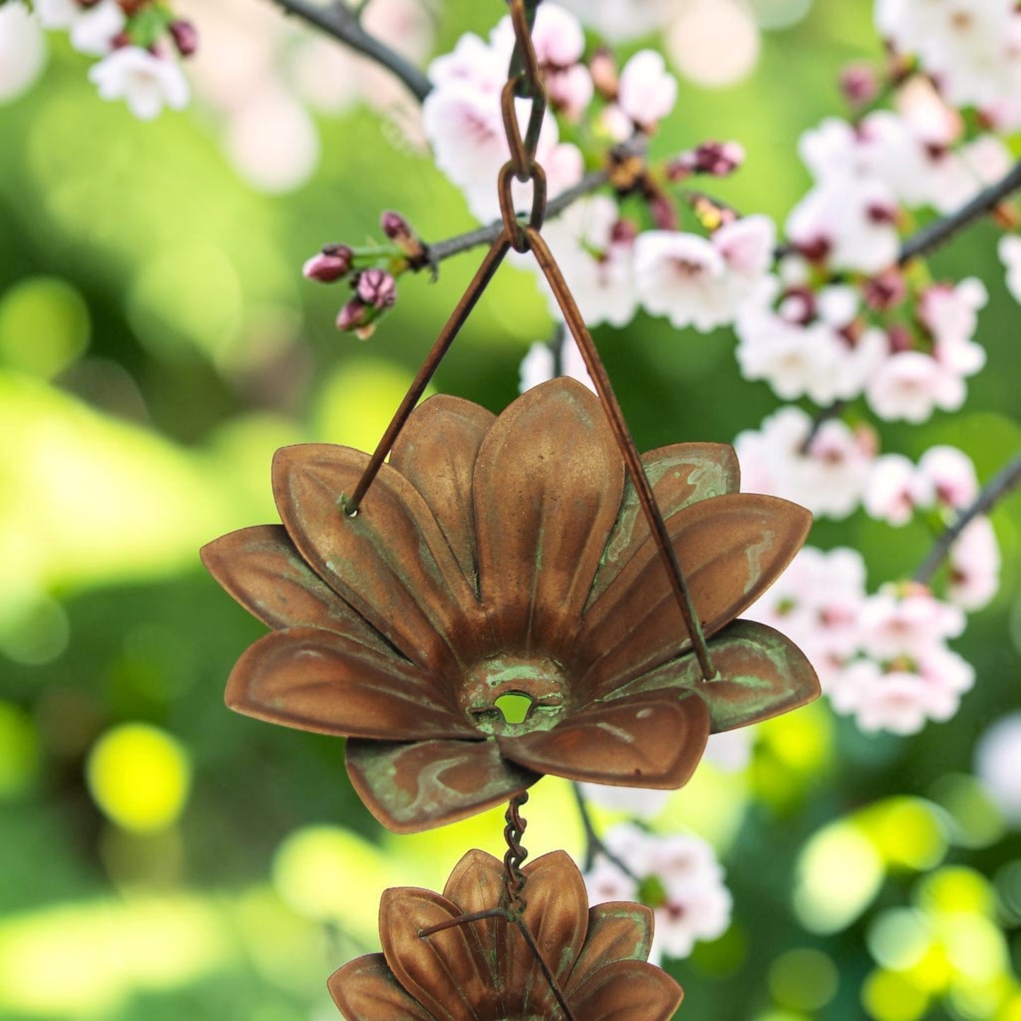 A bronze metal garden wind chime shaped like a lotus flower, hanging from a chain outdoors with blooming pink flowers and green foliage in the background.