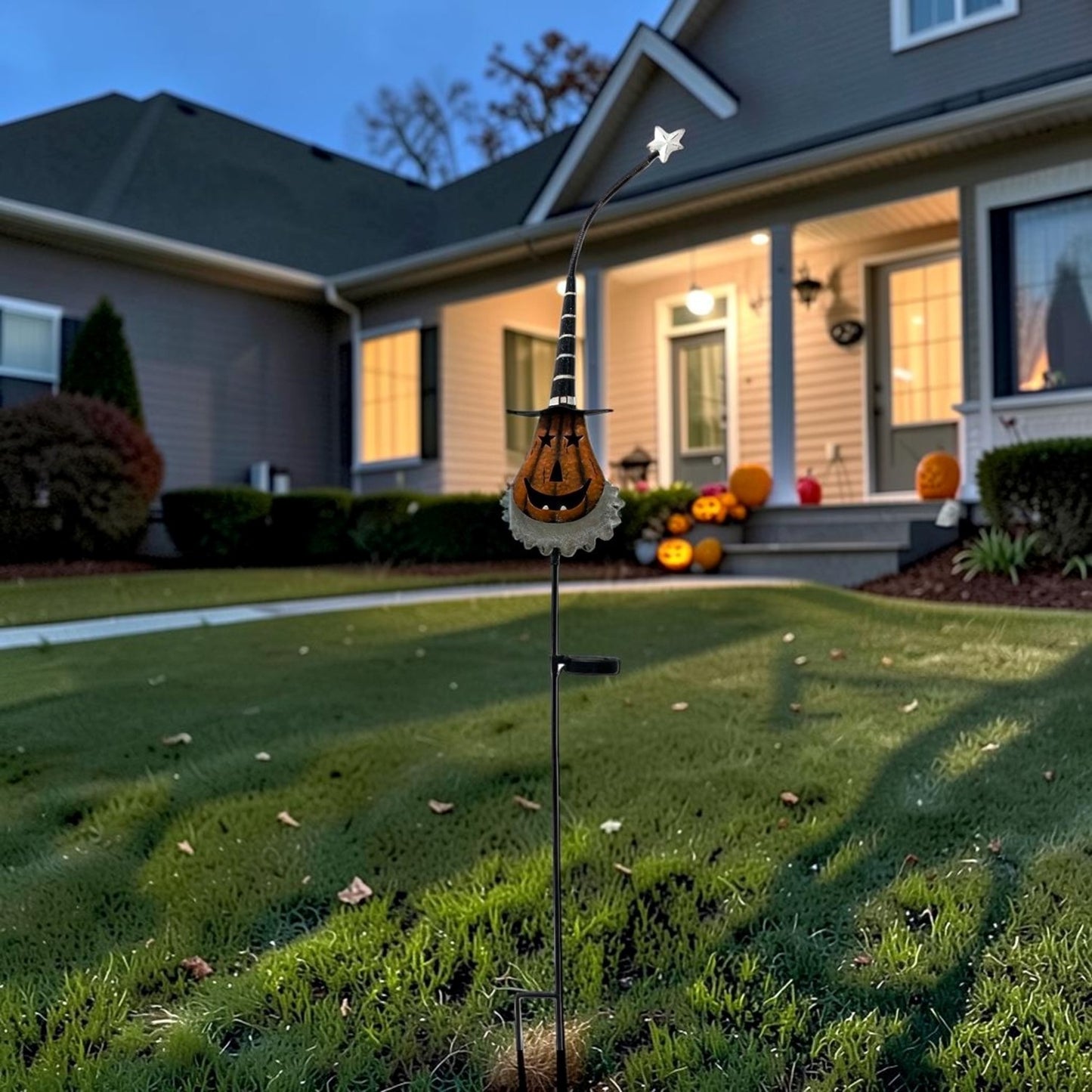 A Halloween yard stake garden ornament featuring a jack-o'-lantern face with a witch hat and a small star on a curved top, displayed in a front yard lawn with a decorated house in the background.