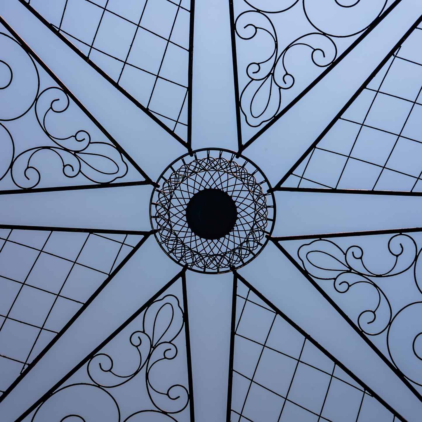 A close-up of a light blue umbrella canopy showing its geometric starburst pattern with black metal framing, diamond grid sections, and elegant scrollwork.