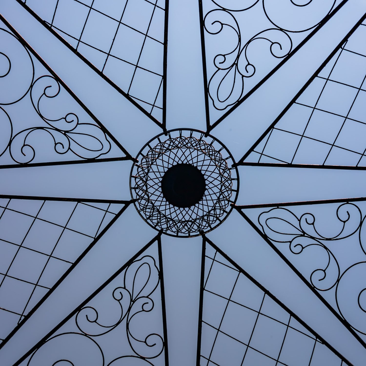 A close-up of a light blue umbrella canopy showing its geometric starburst pattern with black metal framing, diamond grid sections, and elegant scrollwork.