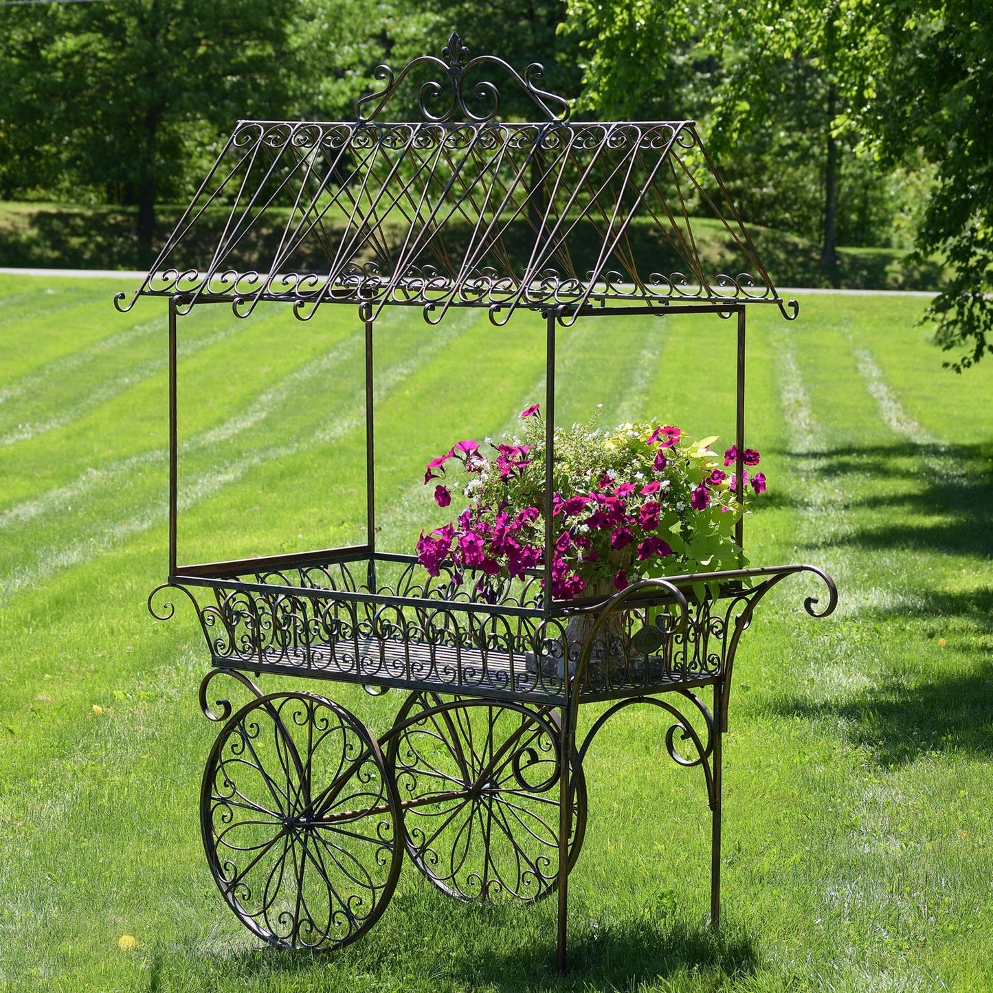 A decorative wrought iron garden cart with a roofed canopy and large spoked wheels, shown outdoors on a green lawn with a potted pink flower arrangement in the basket. Trees and grass are visible in the background.