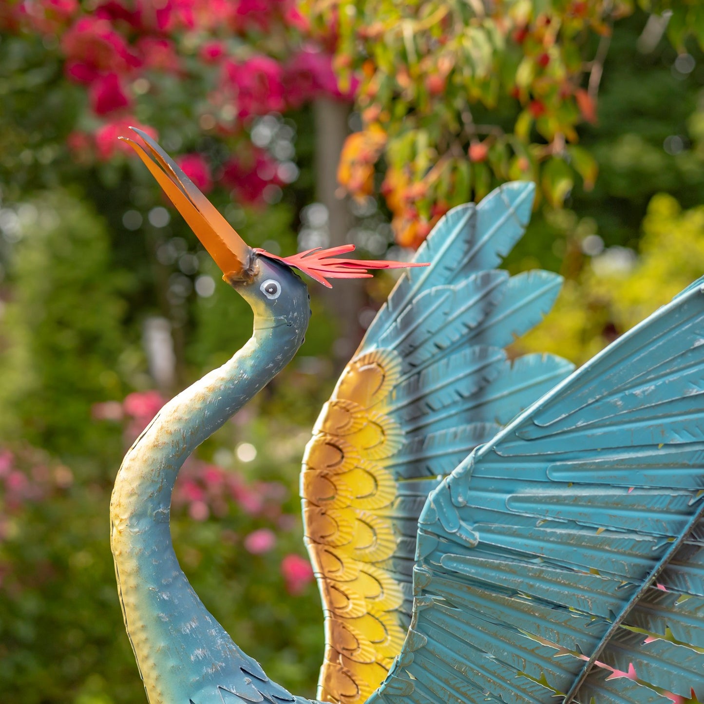 A decorative metal garden statue of a heron with turquoise-blue body and wings, a yellow underwing, and an orange beak, photographed outdoors against a blurred green and pink garden background.