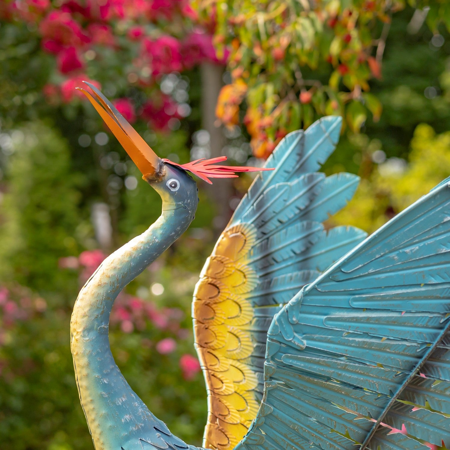 A decorative metal garden statue of a heron with turquoise-blue body and wings, a yellow underwing, and an orange beak, photographed outdoors against a blurred green and pink garden background.