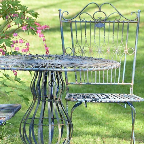 A distressed blue metal outdoor bistro set featuring a round table and two chairs on a green grass lawn, with a flowering plant in the foreground.
