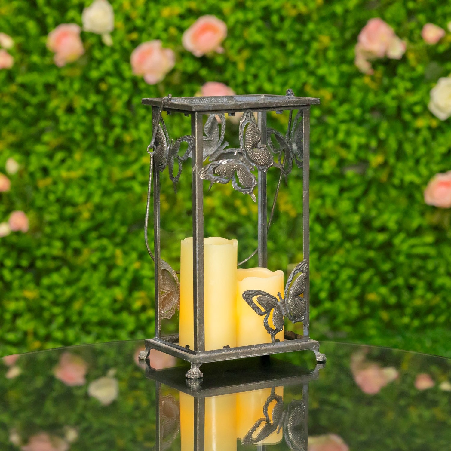 A decorative metal lantern with butterfly details, holding two lit cream pillar candles, set on a reflective surface against a blurred green hedge background with pink flowers.