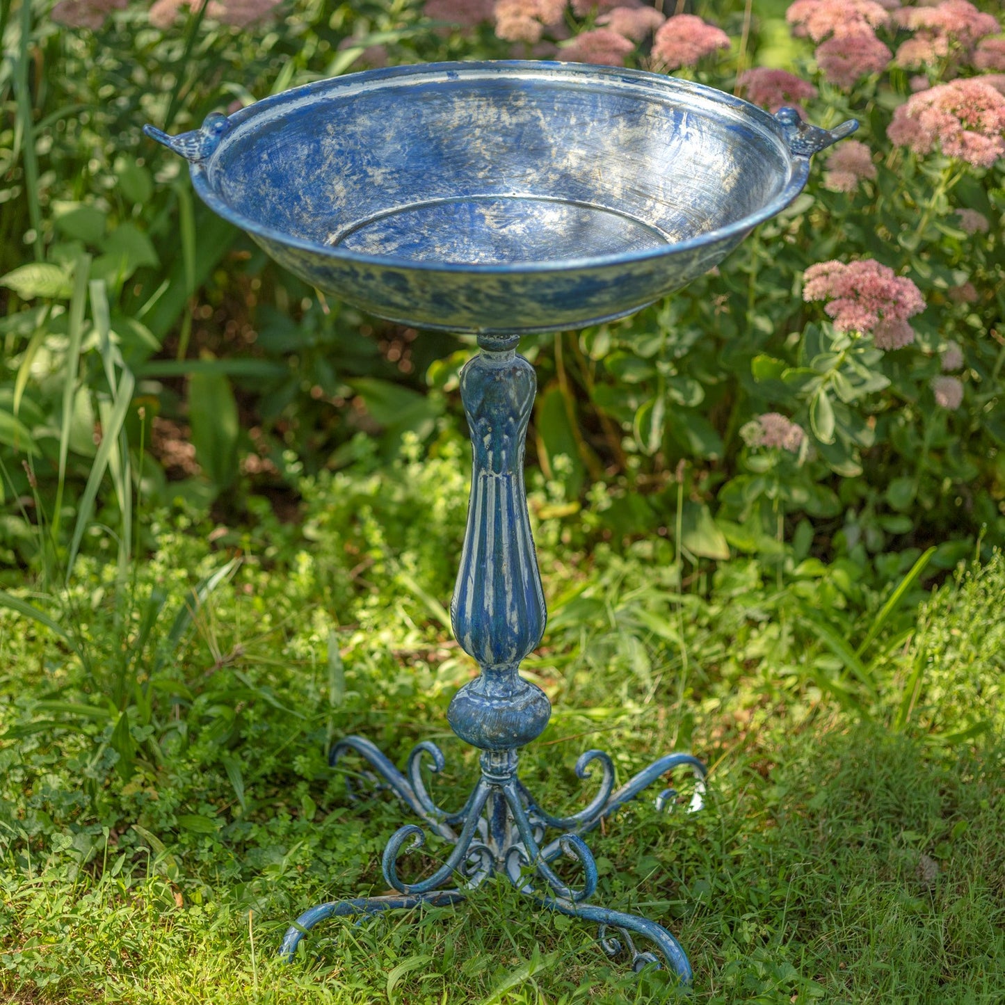 Decorative metal bowl on a stand in a garden setting with flowers and greenery.