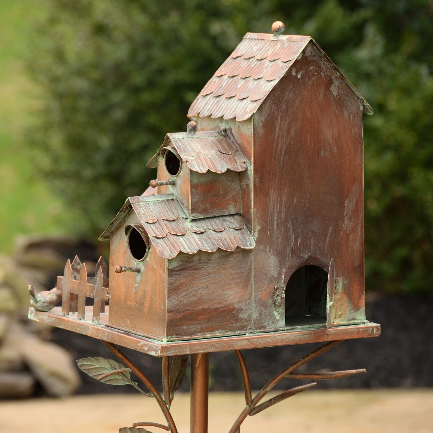 A decorative copper birdhouse with a weathered patina finish, mounted on a metal branch-style stand in an outdoor garden setting.