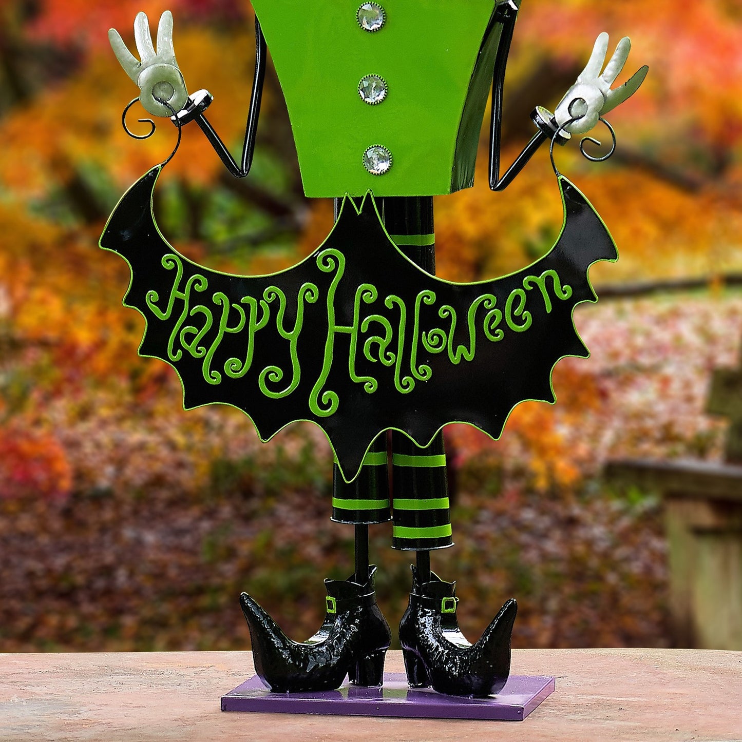 A Halloween metal figurine of a green and black character with white gloves, holding a 'Happy Halloween' sign, standing on a purple base outdoors.