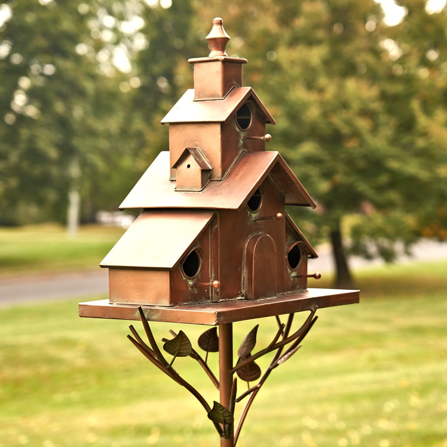 A multi-level copper birdhouse mounted on a decorative metal stake with leaf and branch motifs, photographed outdoors against a blurred green lawn and trees.