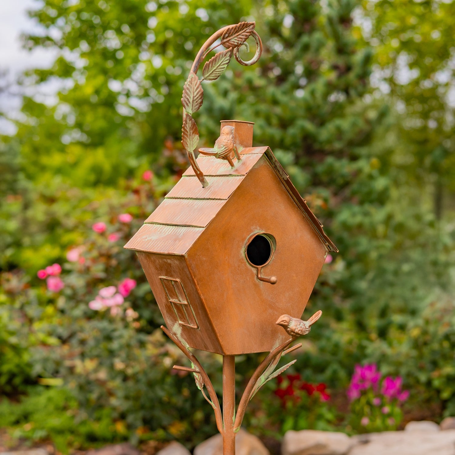 Decorative copper birdhouse with a small bird on top, set against a garden background with flowers and trees