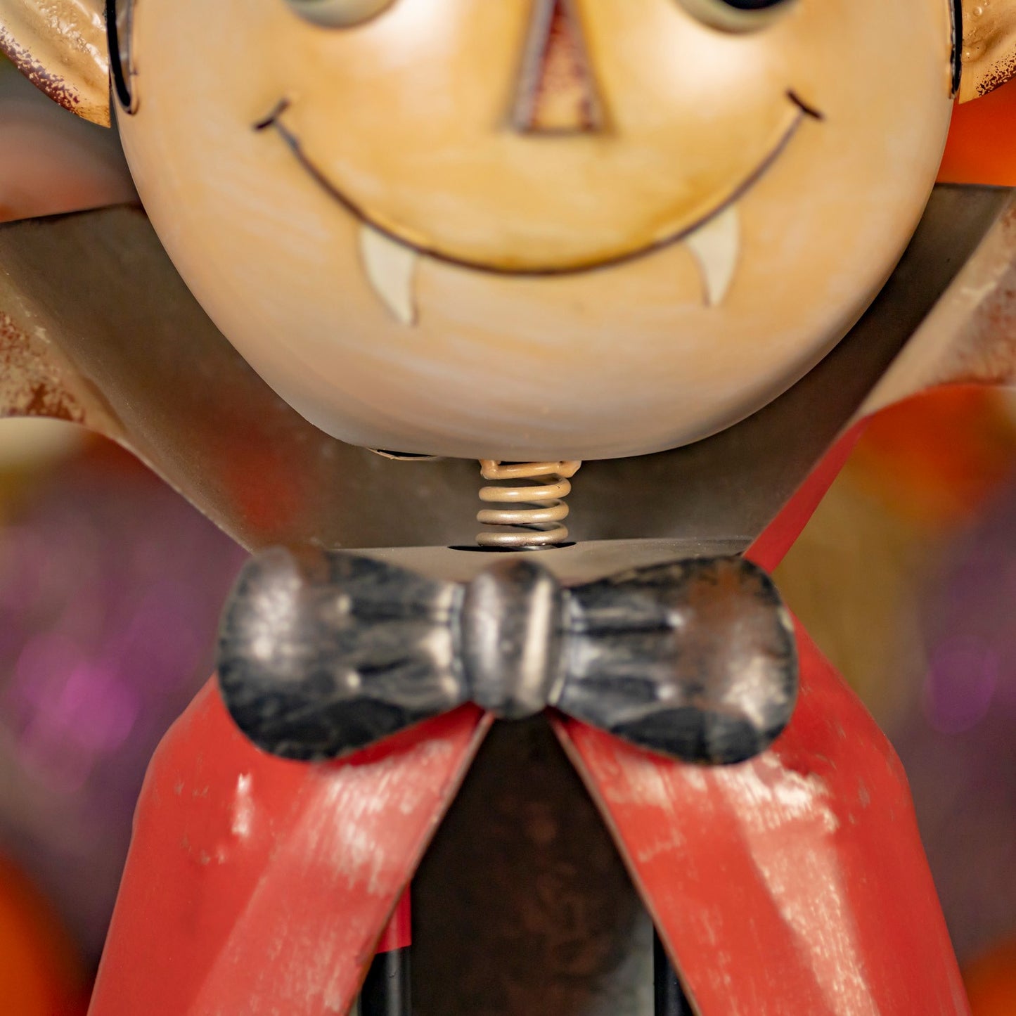 Close-up of a vampire figurine with a round beige face, drawn smile, fangs, dark wings, a black bow tie, and a red cape against a blurred multicolor background.