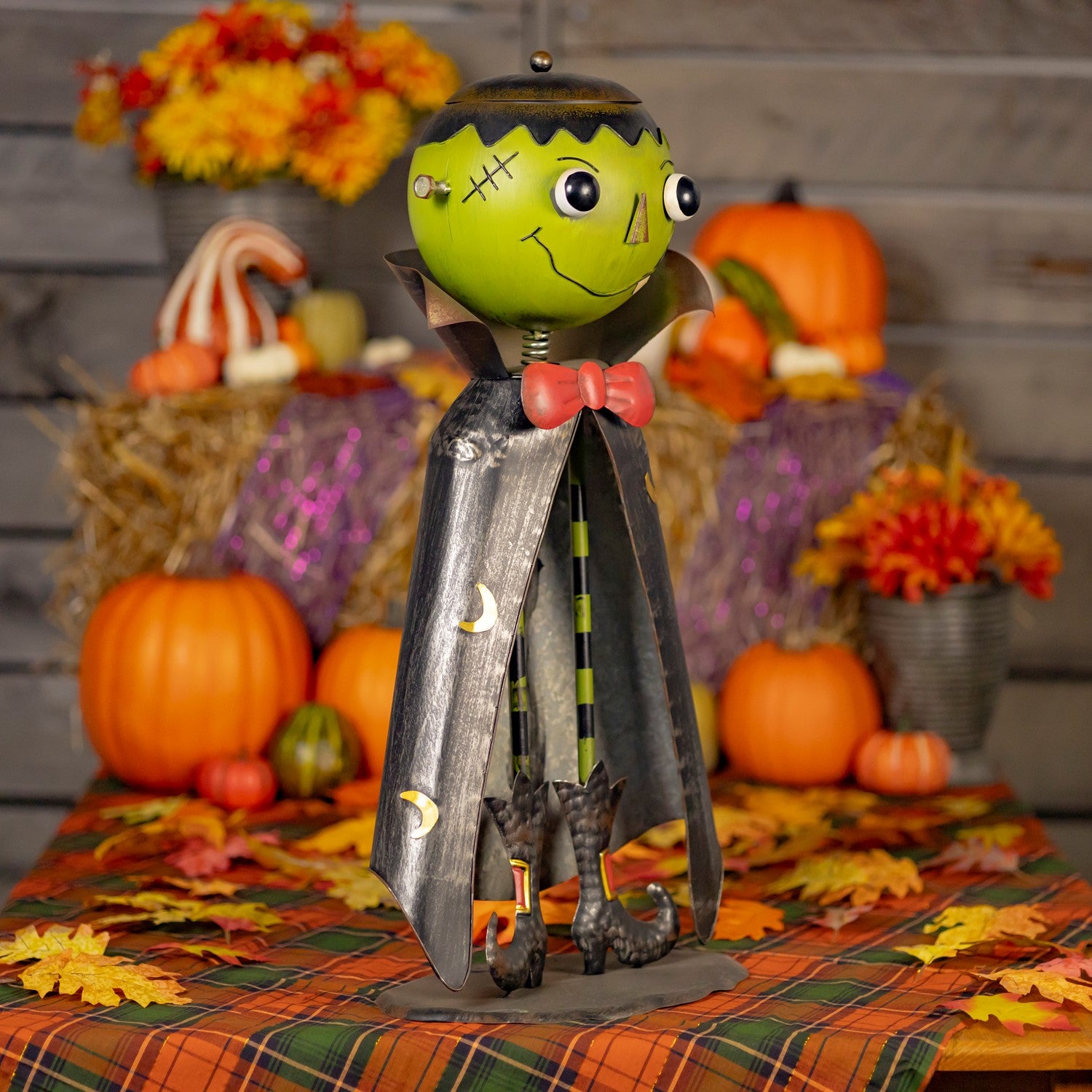 A Halloween decorative metal figure of Frankenstein's monster stands on a plaid autumn tablecloth with pumpkins, hay bales, and fall flowers in the background.