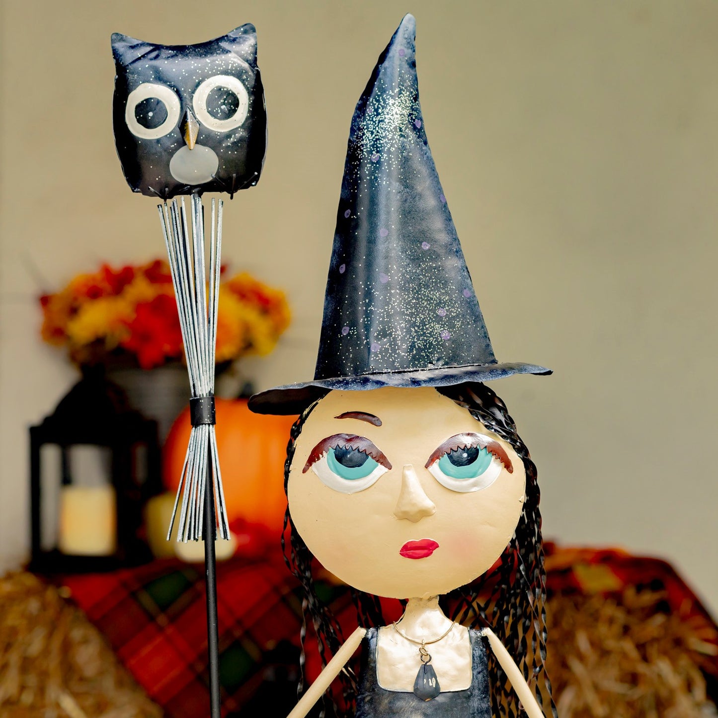 A handmade witch doll with a large round face, blue eyes, and a tall glittery black hat, standing next to a black owl-shaped candle holder on a stick. The background features a soft-focus autumn setting with a pumpkin, plaid fabric, and hay.