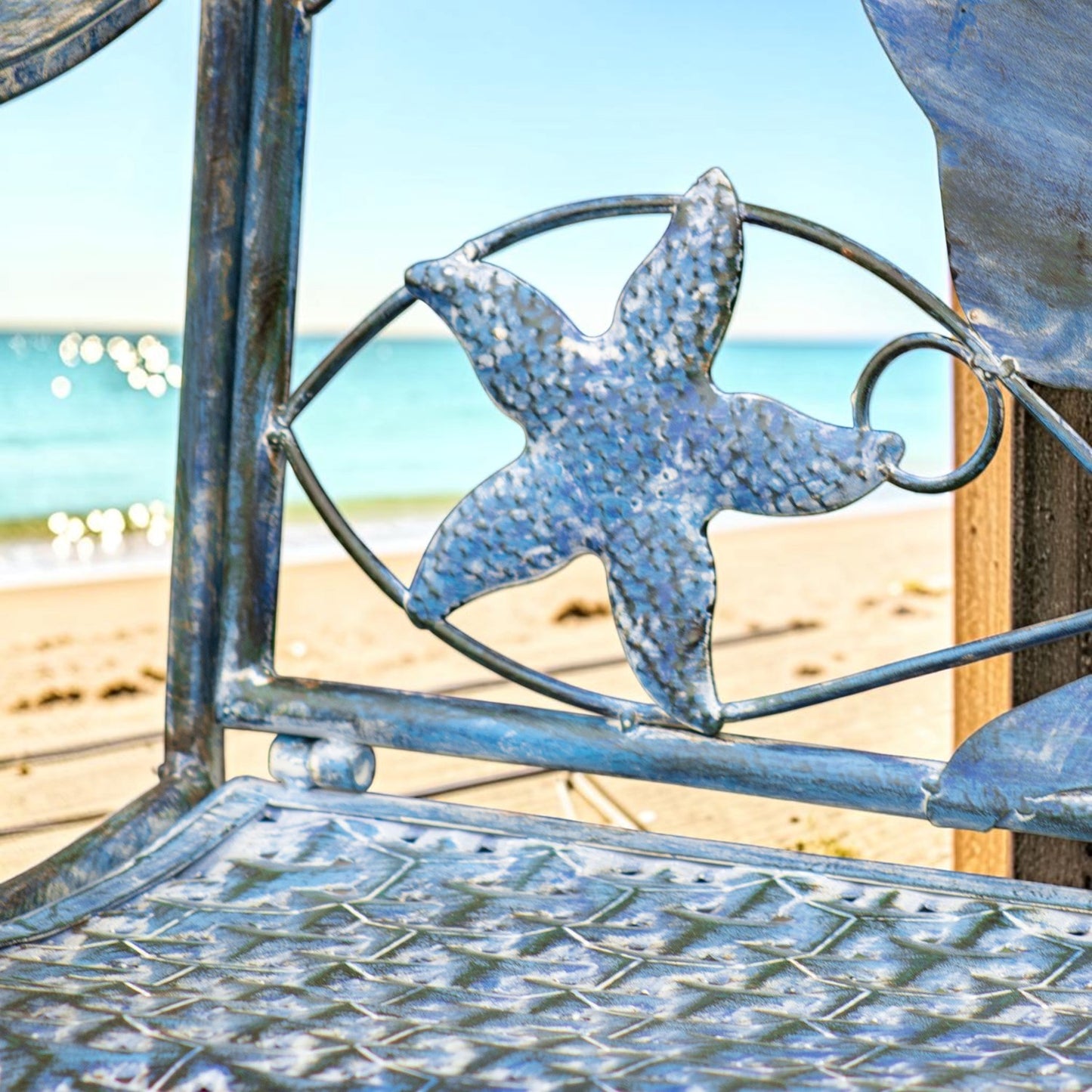 A close-up of a weathered blue metal beach chair with a hammered starfish design on the backrest, set against a blurred beach and ocean background.