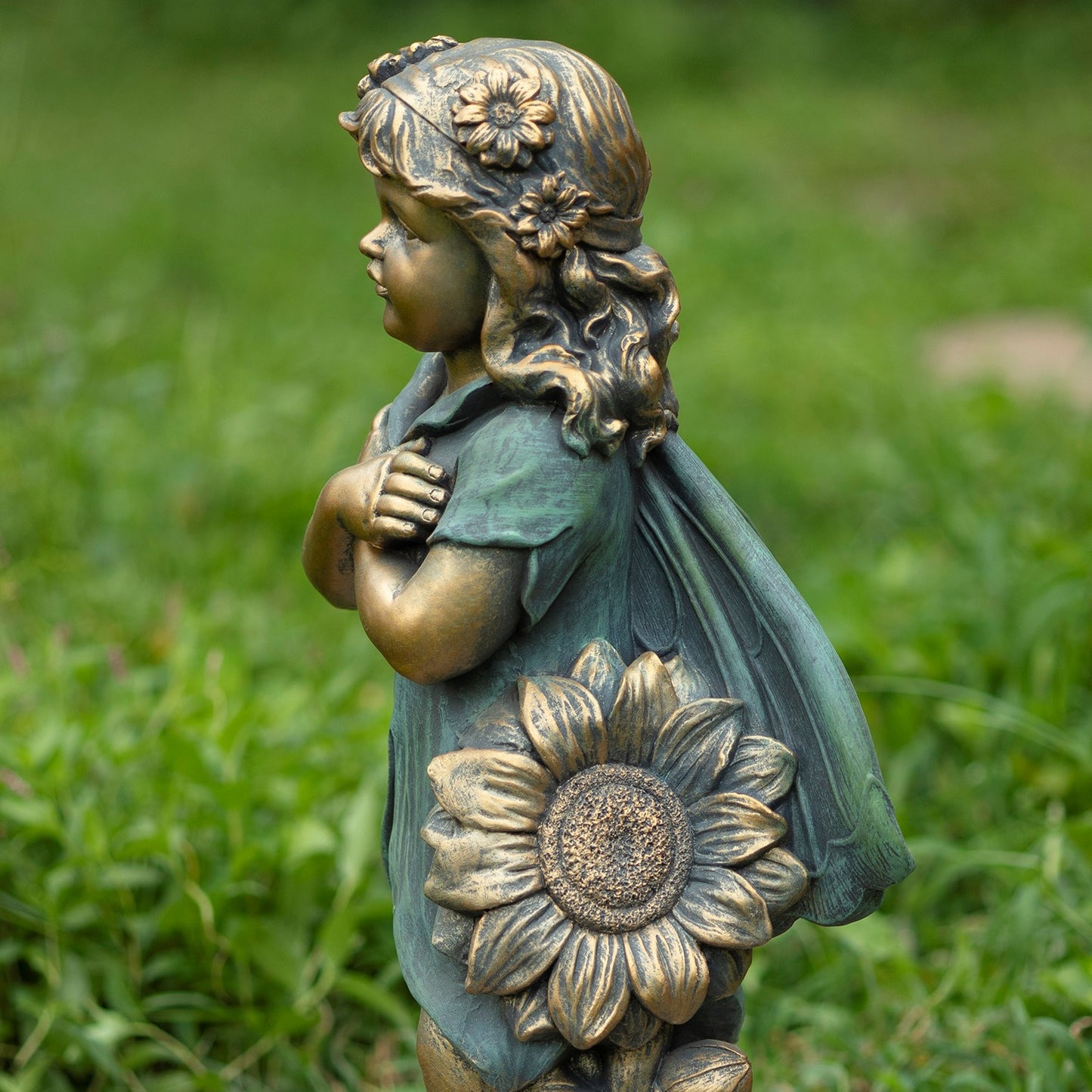 A bronze-toned garden statue of a young girl with a flower crown and a flowing cape, holding a large sunflower, photographed outdoors on a blurred green lawn.