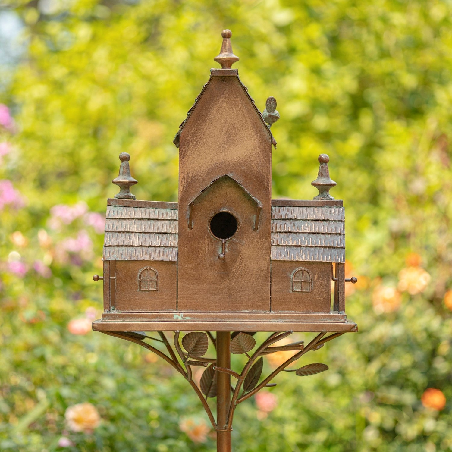 A decorative copper birdhouse with a miniature cottage design, featuring a central round bird hole, shingled roof details, and a leafy metal stand, set against a blurred green garden background.
