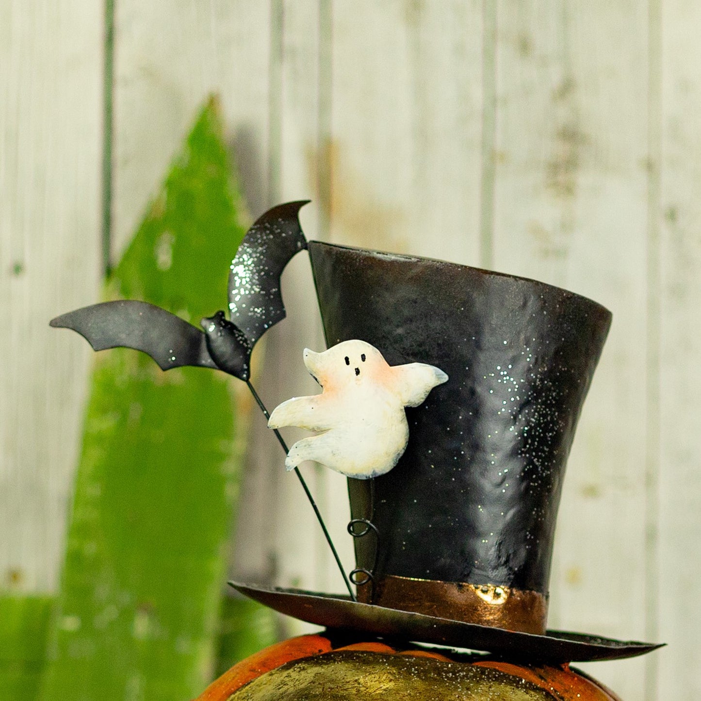 A Halloween decoration featuring a black top hat with silver glitter, a small white ghost, and a black bat on a stand, displayed on top of a pumpkin against a rustic wood plank background.