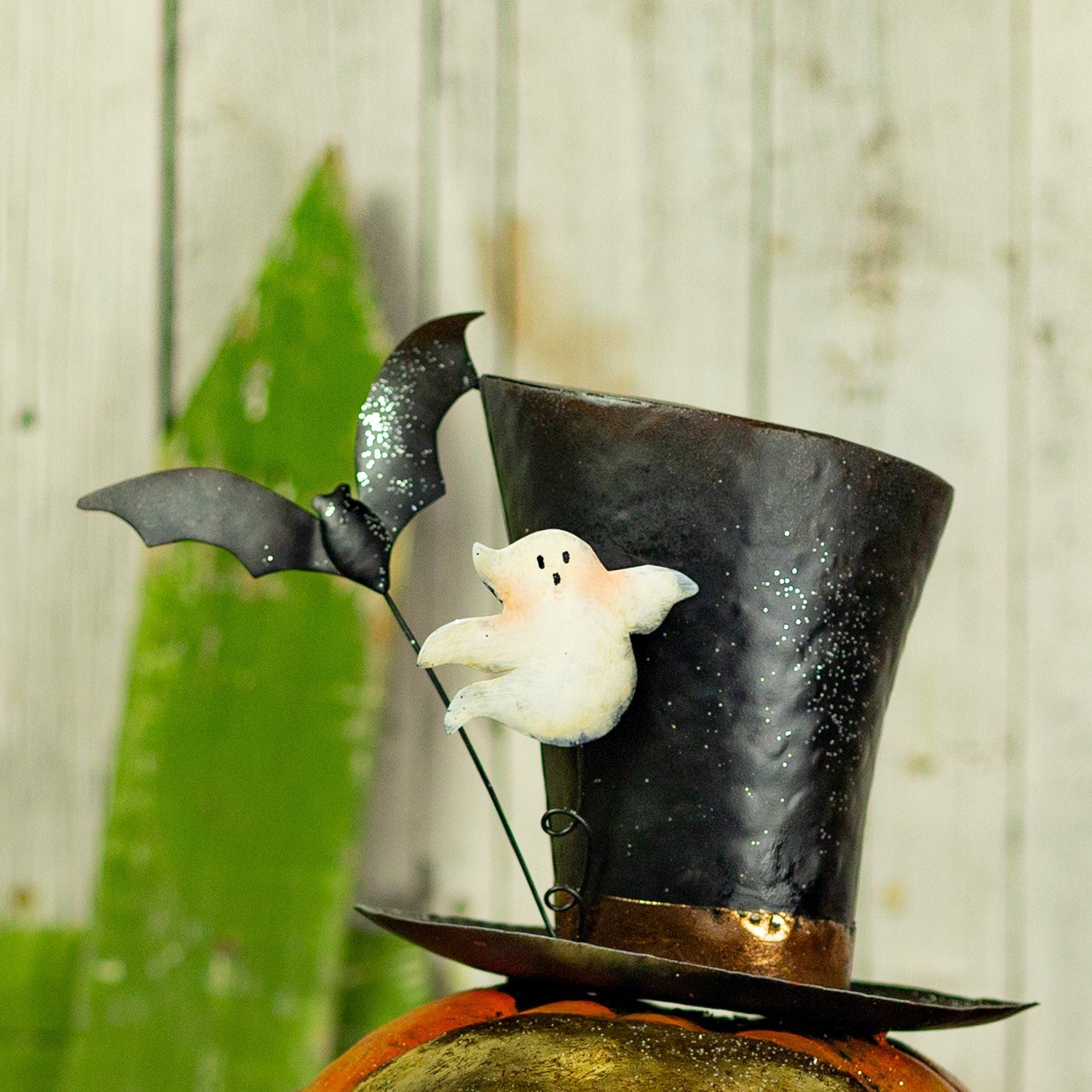 A Halloween decoration featuring a black top hat with silver glitter, a small white ghost, and a black bat on a stand, displayed on top of a pumpkin against a rustic wood plank background.