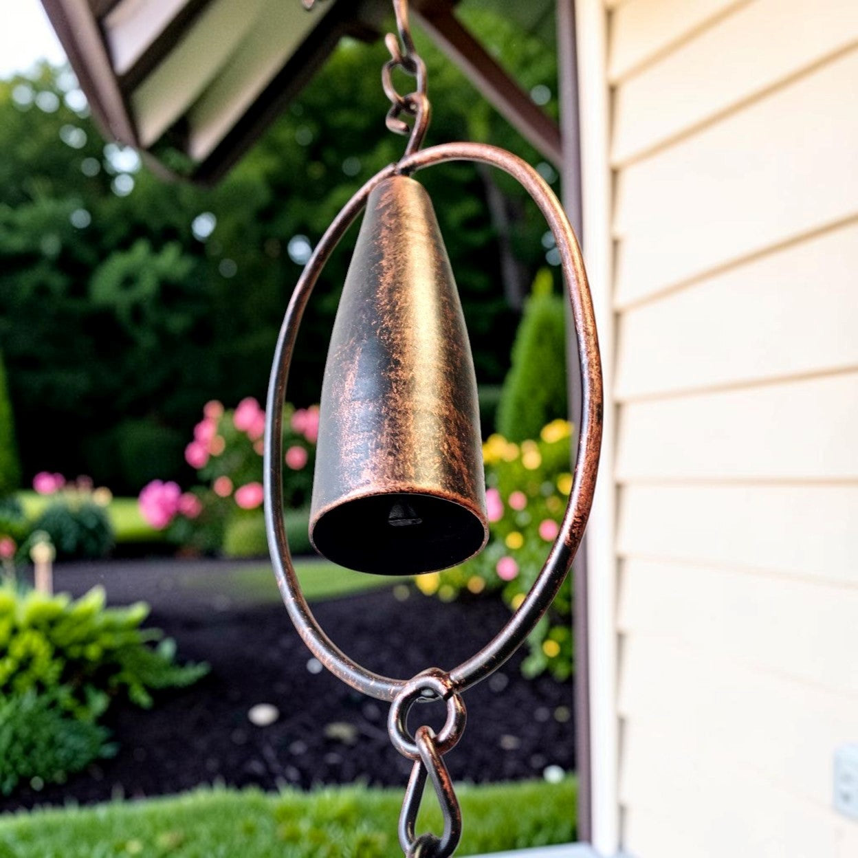 A rustic bronze metal wind chime hanging outdoors in front of a house, featuring a conical bell suspended inside a large circular hoop with a hanging chain.