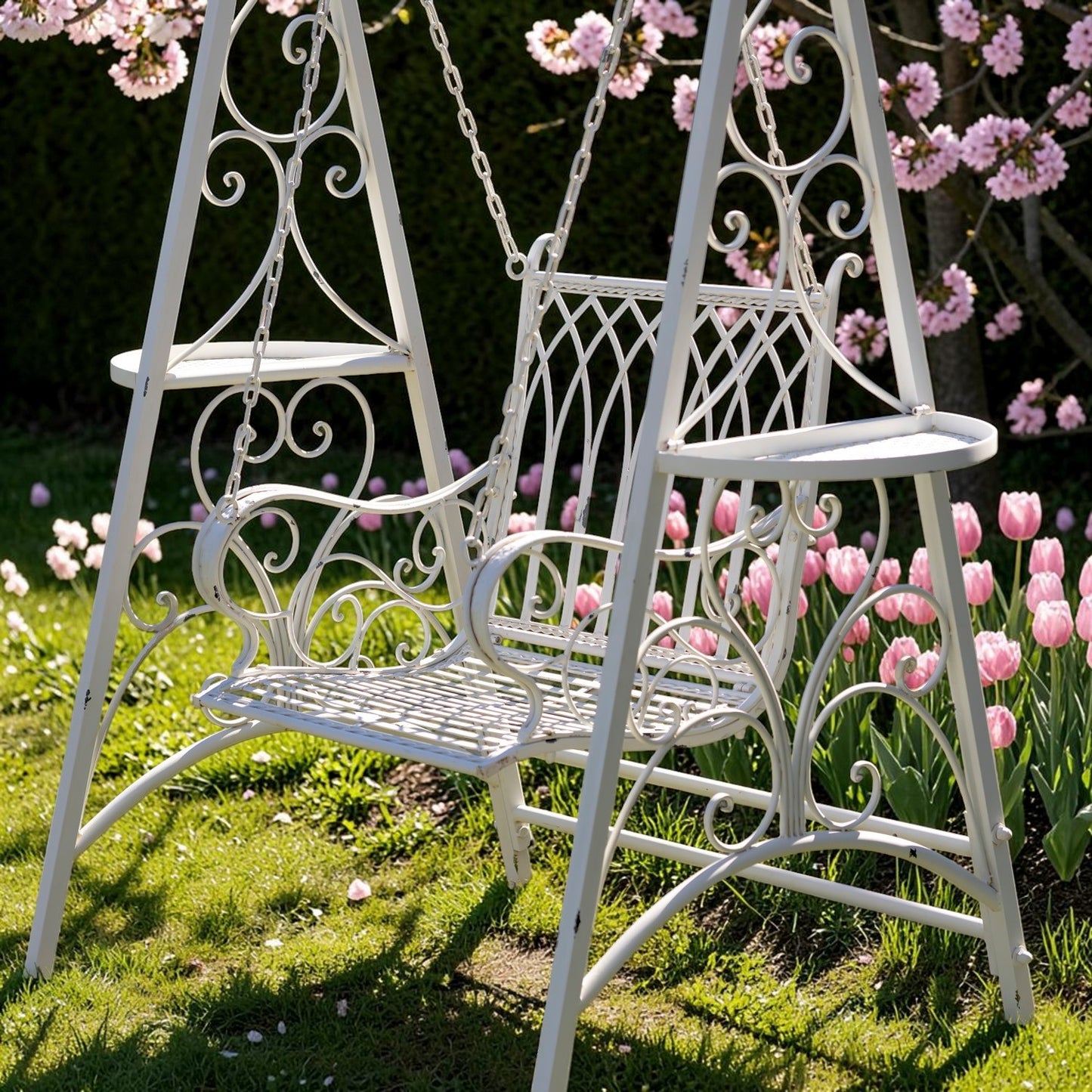 A white wrought-iron garden swing set with a single hanging swing seat, shown outdoors on grass with pink tulips and blossoms in the background.