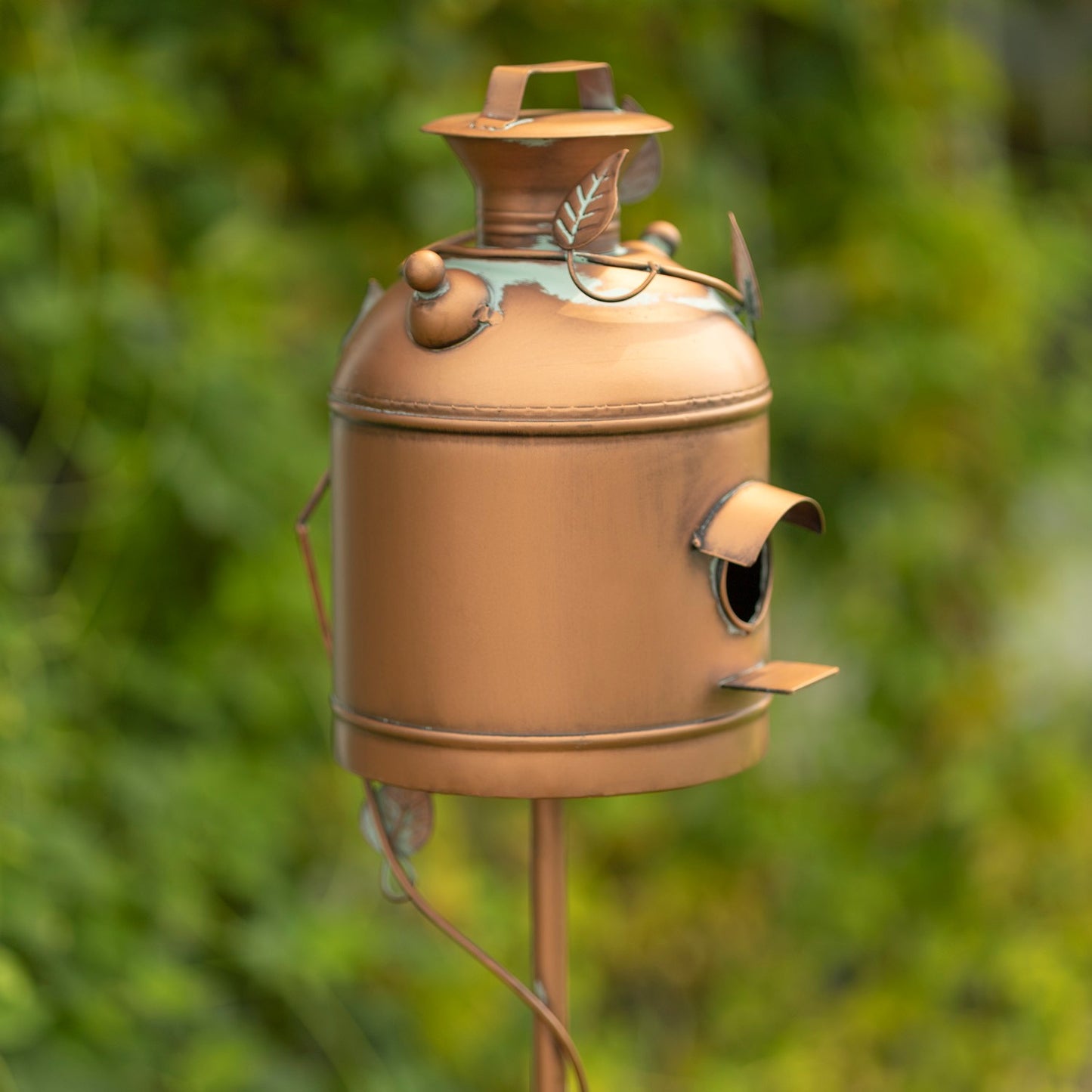 A rustic copper-colored metal birdhouse shaped like a vintage milk canister, mounted on a stake against a blurred green garden background.