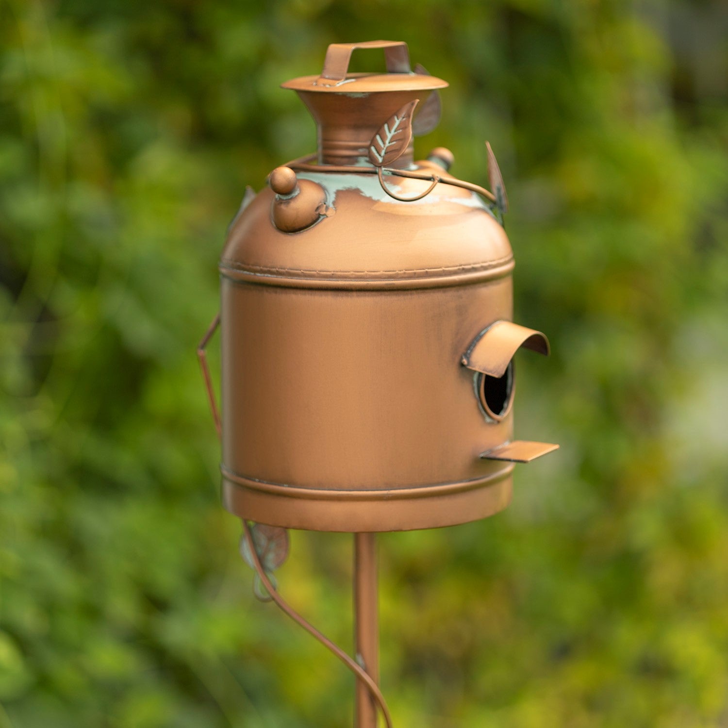 A rustic copper-colored metal birdhouse shaped like a vintage milk canister, mounted on a stake against a blurred green garden background.
