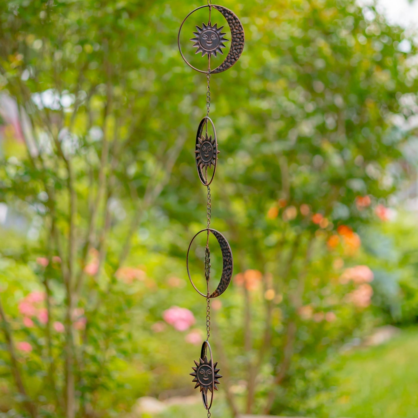 A hanging metal garden ornament featuring three moon and sun phases connected by chains, photographed outdoors against a blurred green garden background.