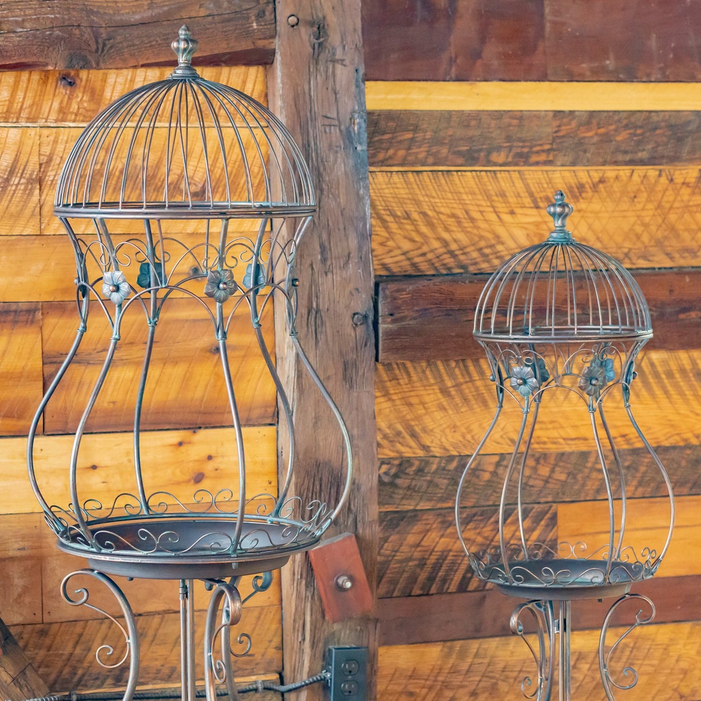 Two vintage-style metal bird baths with domed wire cages and tray bases, displayed against a rustic wooden plank wall.