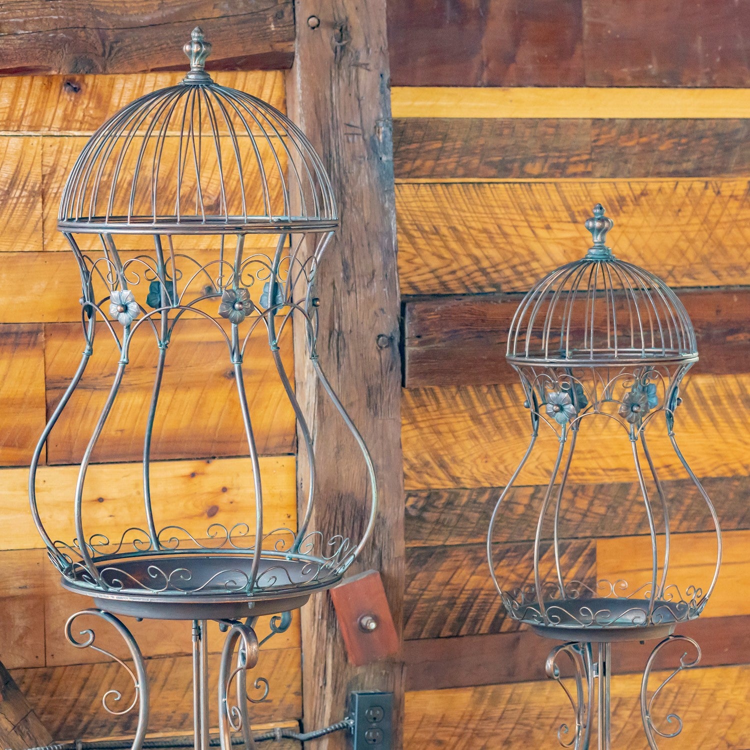 Two vintage-style metal bird baths with domed wire cages and tray bases, displayed against a rustic wooden plank wall.