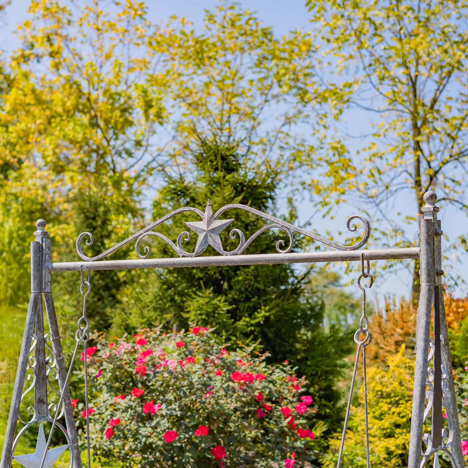 A decorative metal swing gate or garden arch in a weathered silver finish, set outdoors in a garden with green trees and a bush of bright pink flowers under a blue sky.