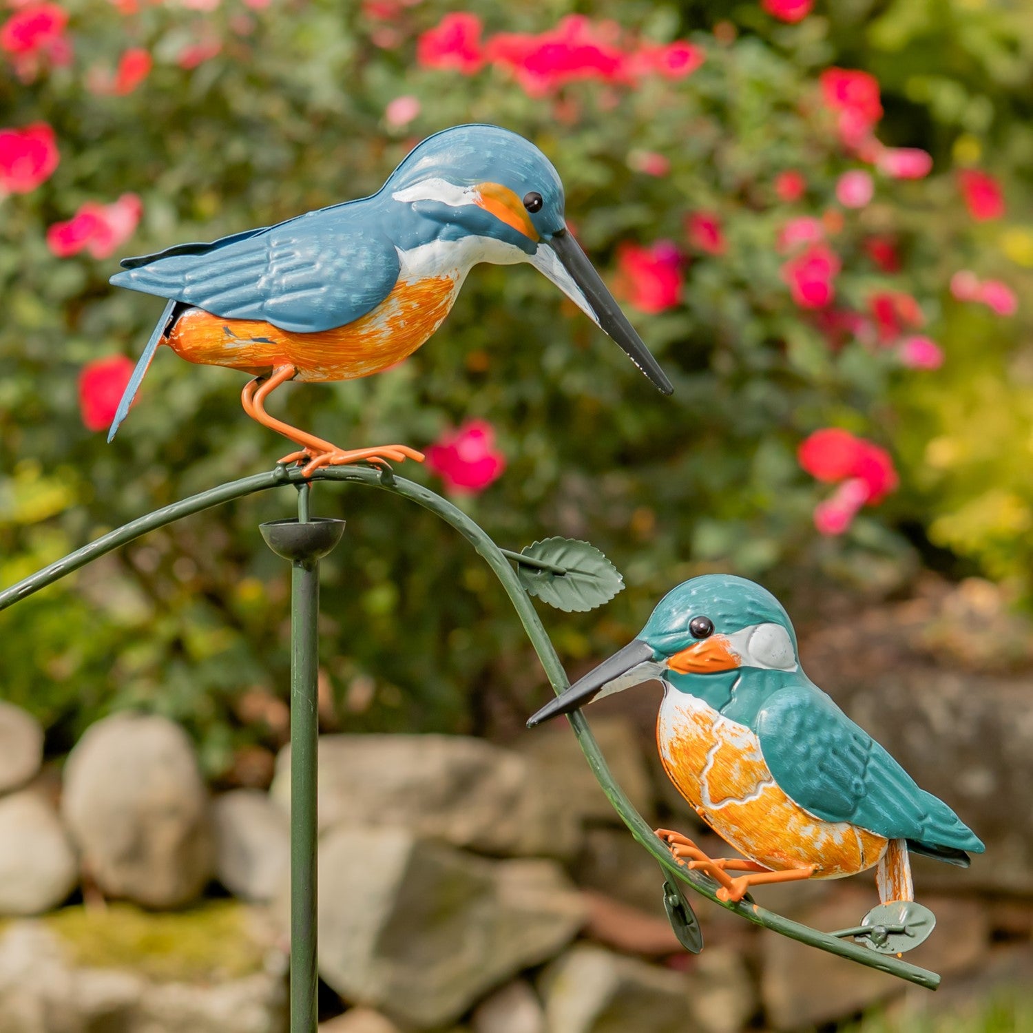 Two decorative kingfisher bird garden statues in blue, orange, and white, mounted on a curved green metal stake in a garden setting with blurred pink flowers and greenery.