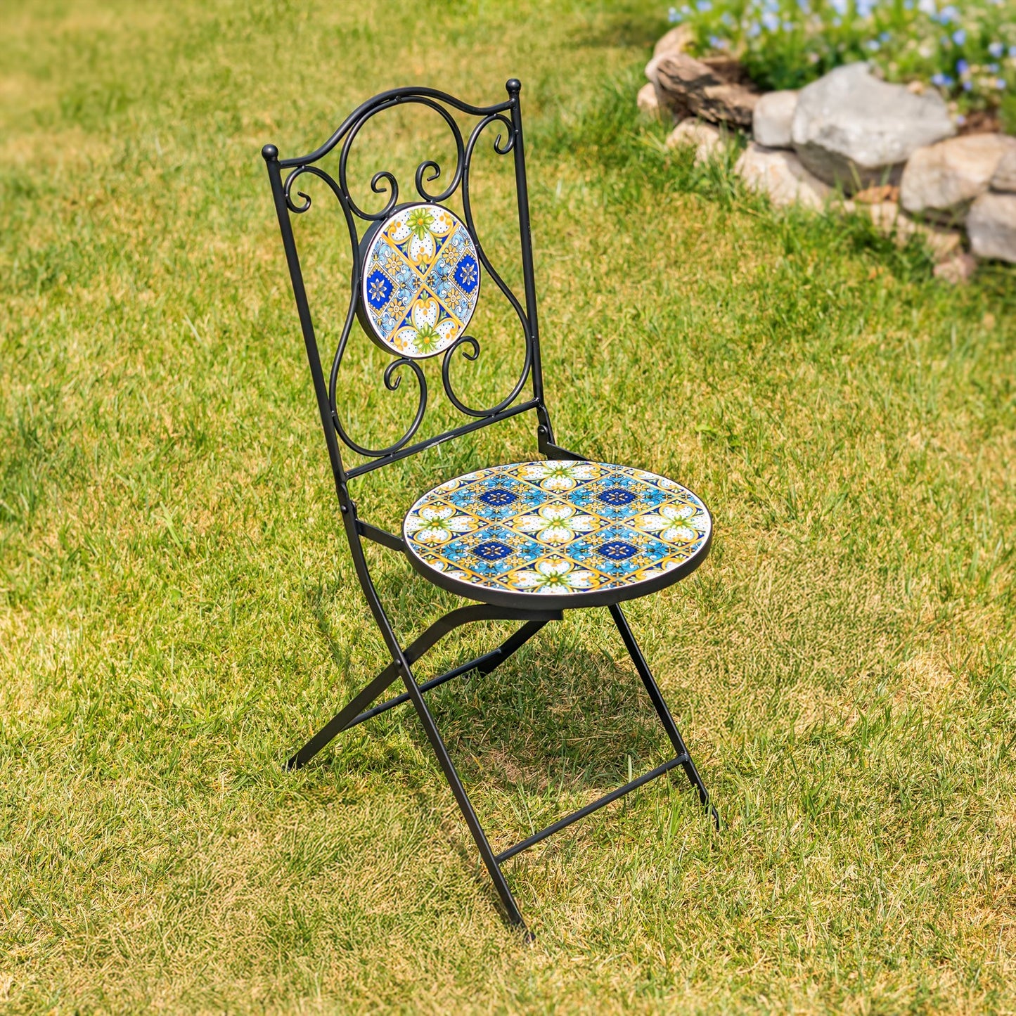 A black metal folding garden chair with a round mosaic tile seat and a matching oval tile backrest, placed on a green grass lawn with a stone garden bed in the background.