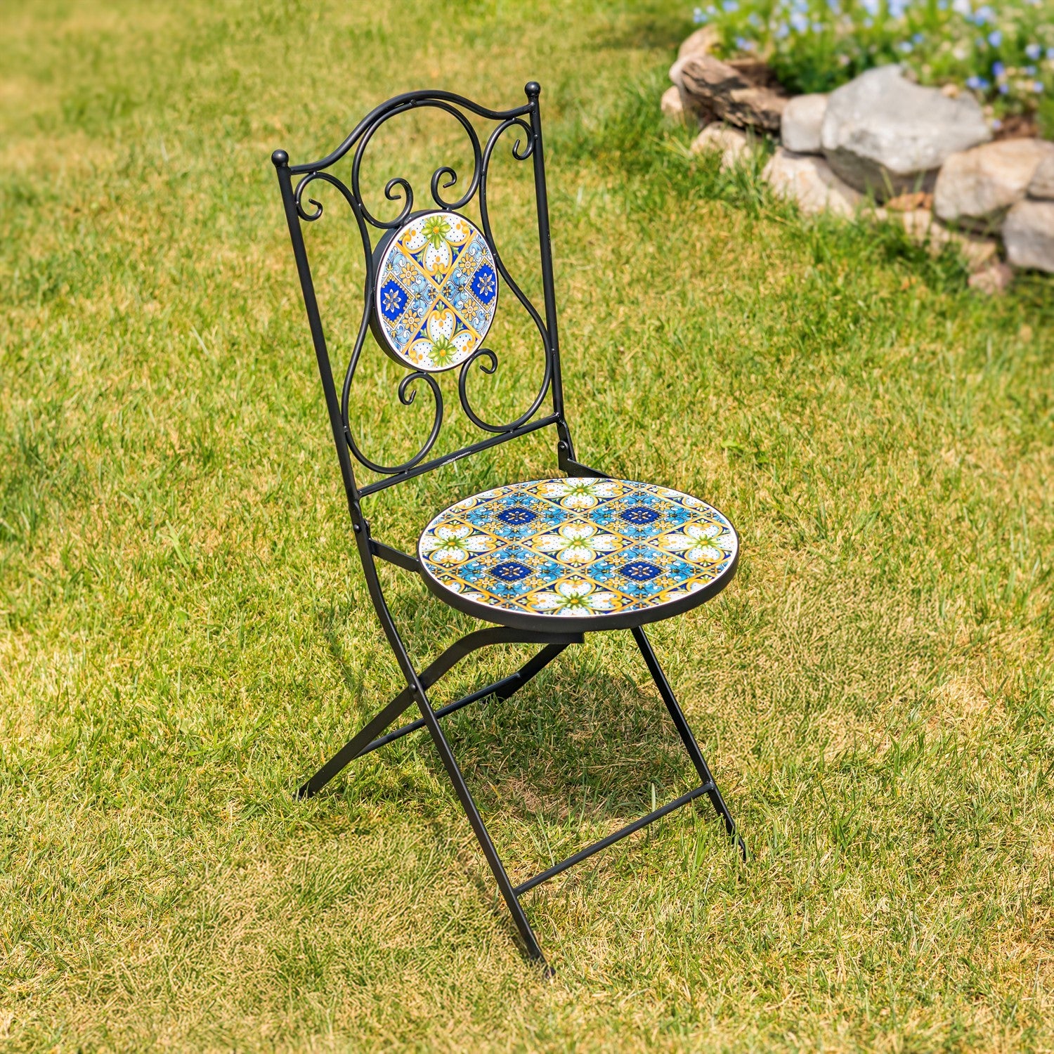 A black metal folding garden chair with a round mosaic tile seat and a matching oval tile backrest, placed on a green grass lawn with a stone garden bed in the background.