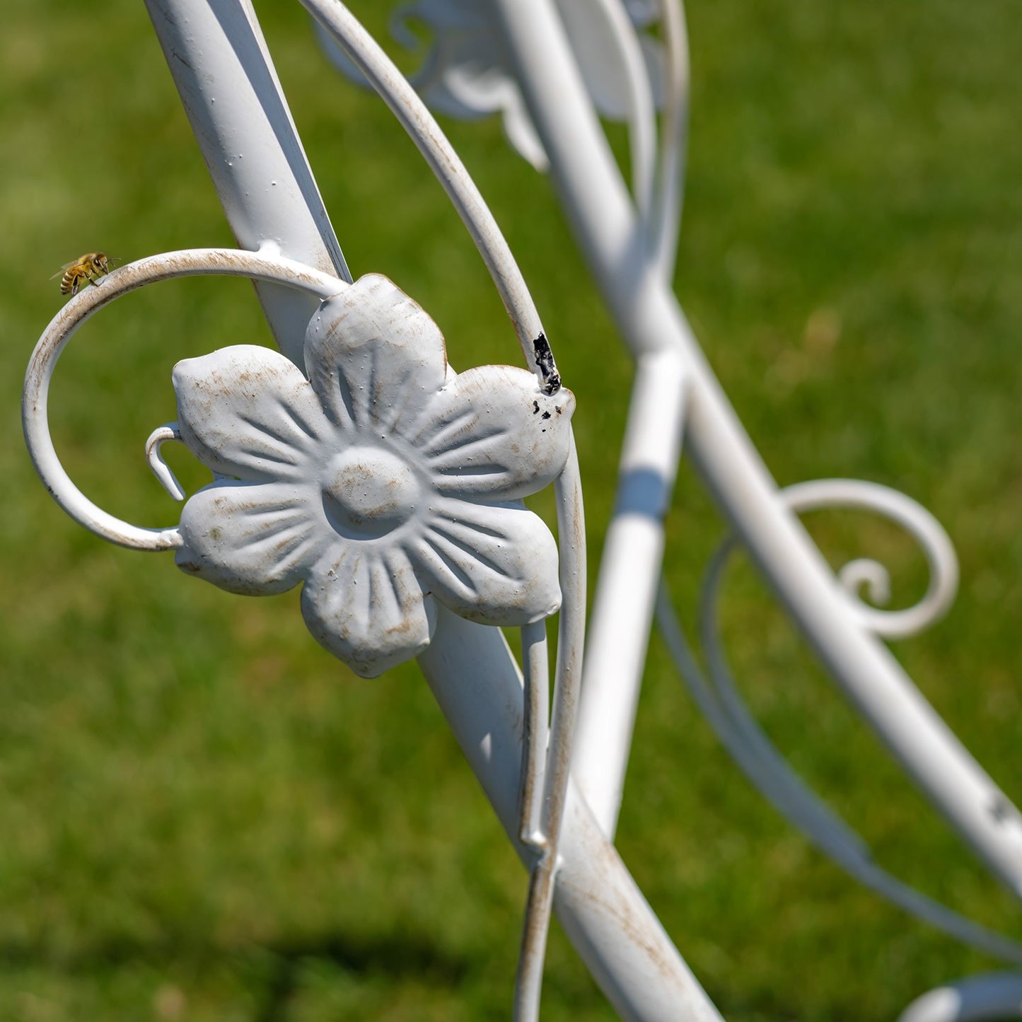 A close-up of a white metal garden bench or chair frame with a decorative flower detail, photographed outdoors against a blurred green grass background.