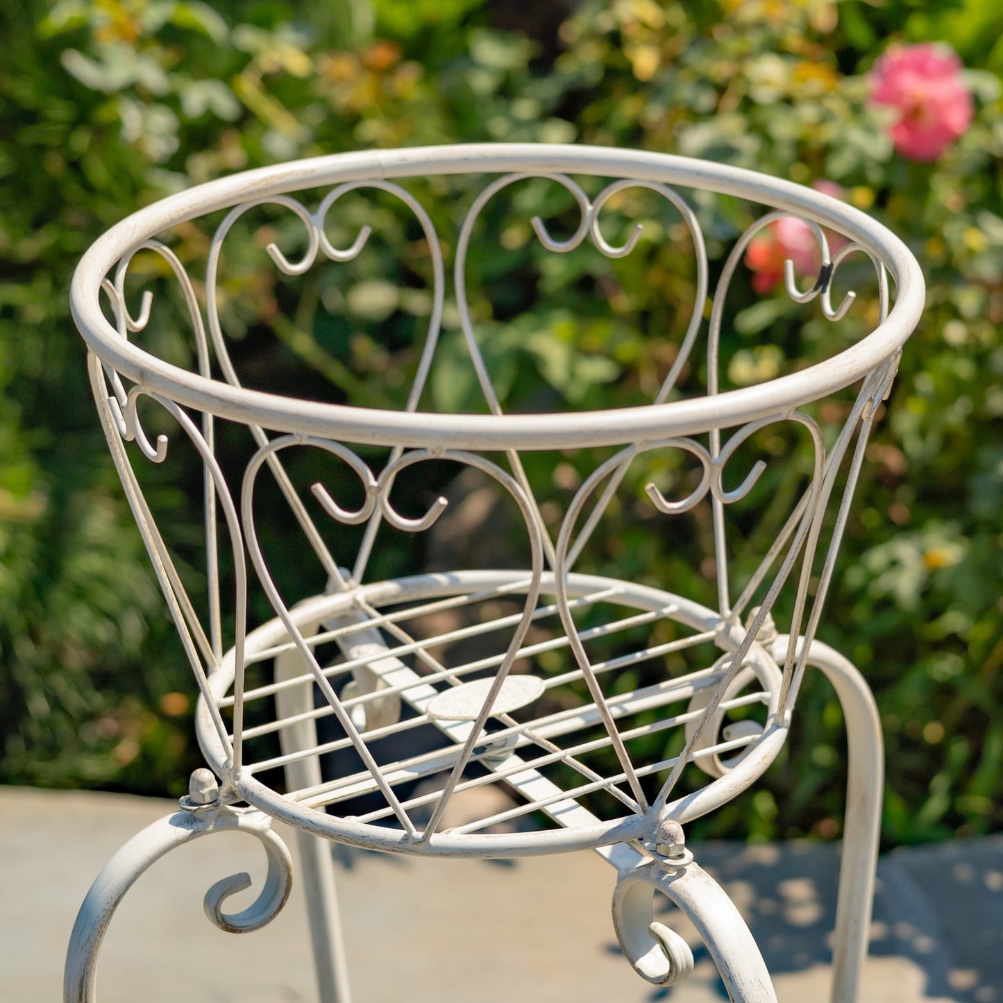 A white metal garden planter basket with decorative scrollwork, shown outdoors on a patio with a blurred green garden background.