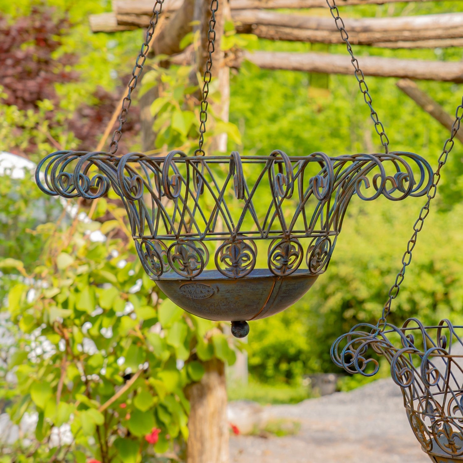 A dark metal hanging planter basket with decorative scrollwork and a bowl-shaped base, suspended by chains in an outdoor garden setting with blurred green foliage and wooden posts.