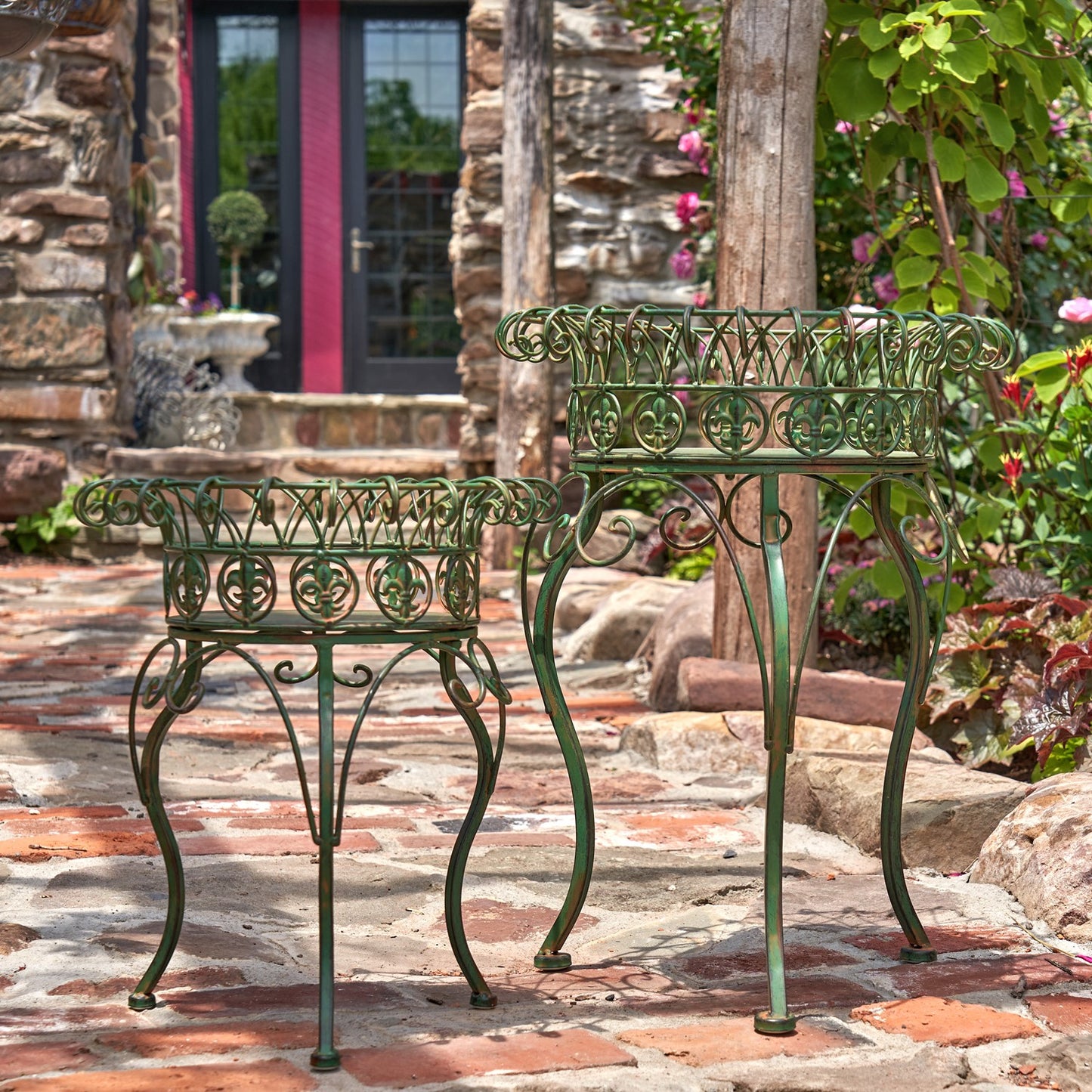 Two green wrought iron garden tables with ornate scrollwork tops and curved legs, displayed on a stone patio in front of a rustic stone house.