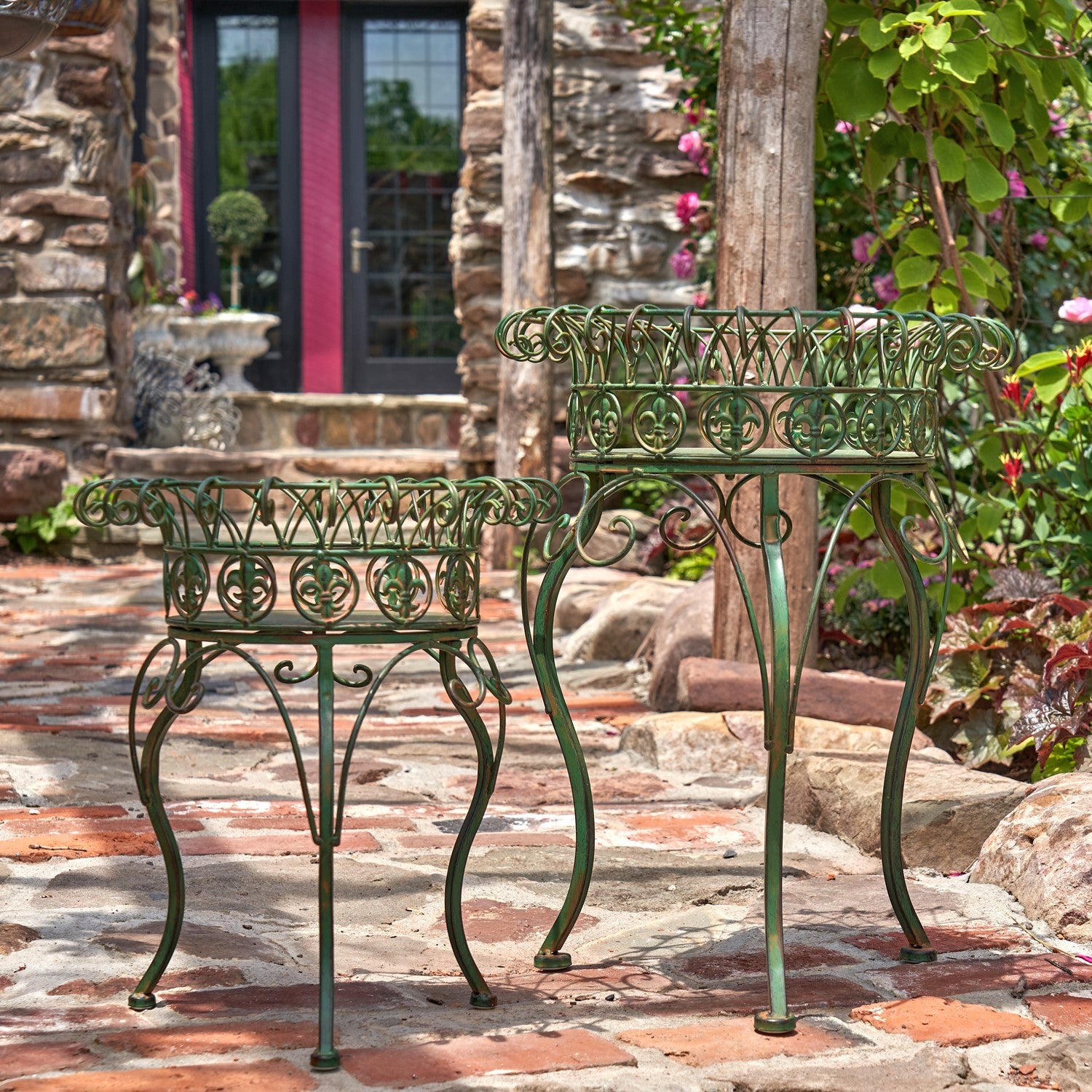 Two green wrought iron garden tables with ornate scrollwork tops and curved legs, displayed on a stone patio in front of a rustic stone house.