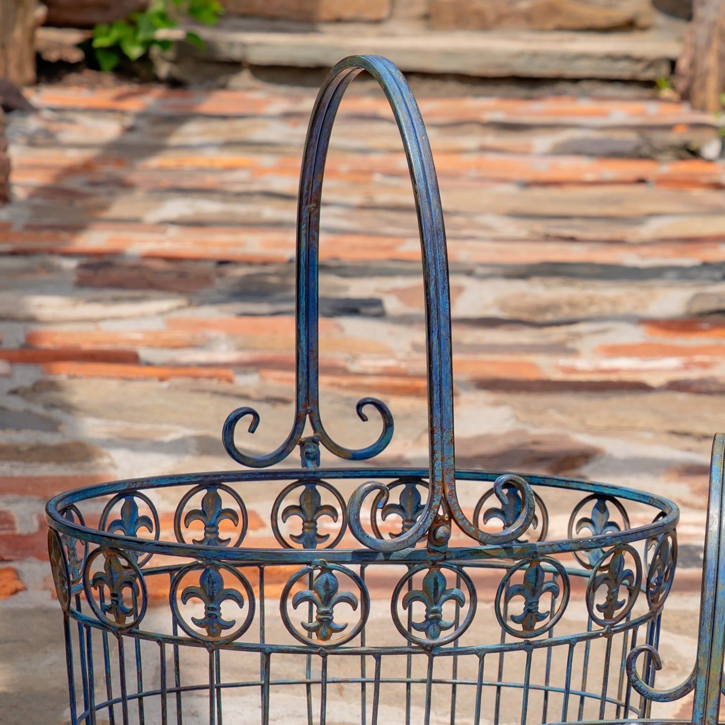 A dark blue wrought iron garden basket with a decorative fleur-de-lis pattern and a tall arched handle, shown outdoors on a stone patio.
