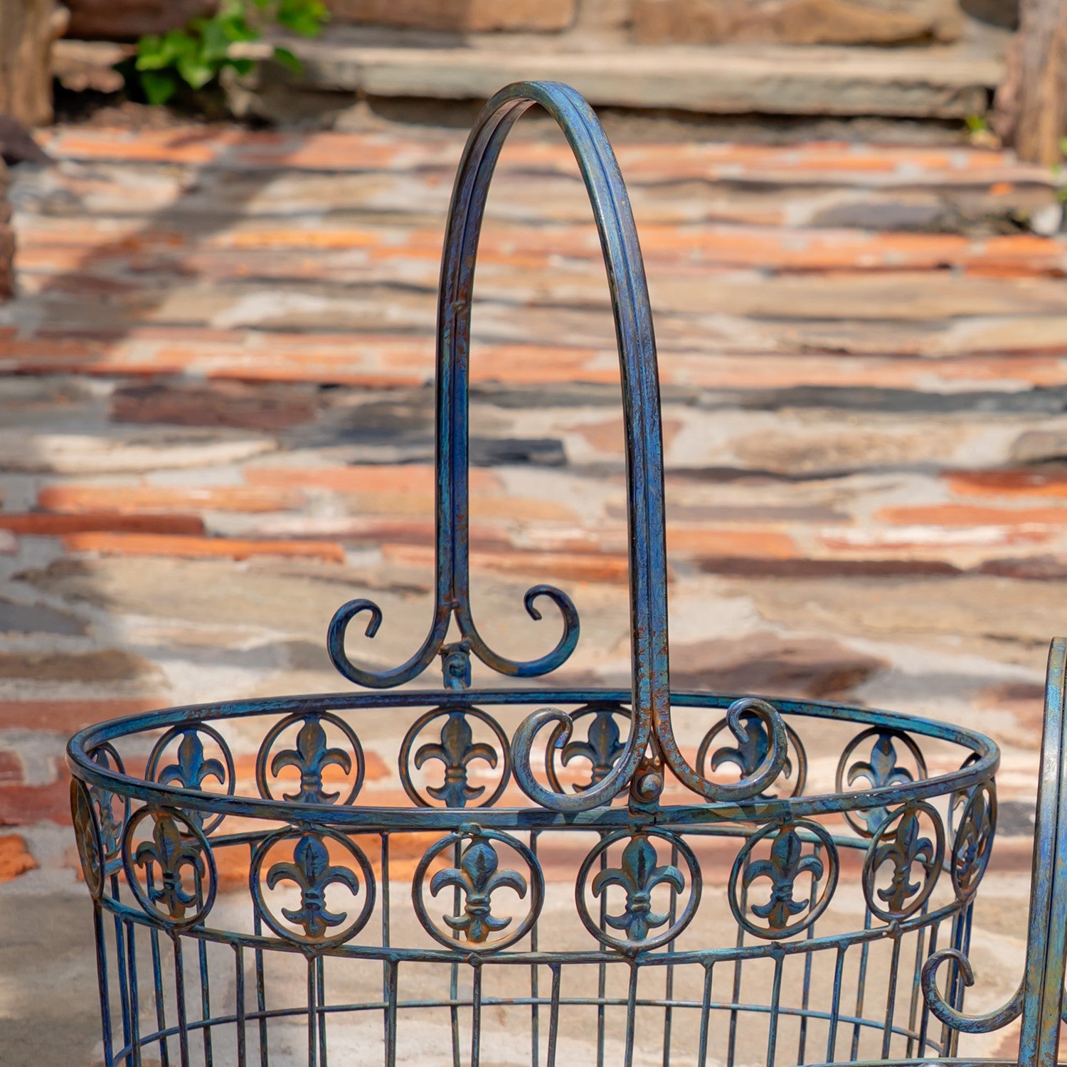 A dark blue wrought iron garden basket with a decorative fleur-de-lis pattern and a tall arched handle, shown outdoors on a stone patio.