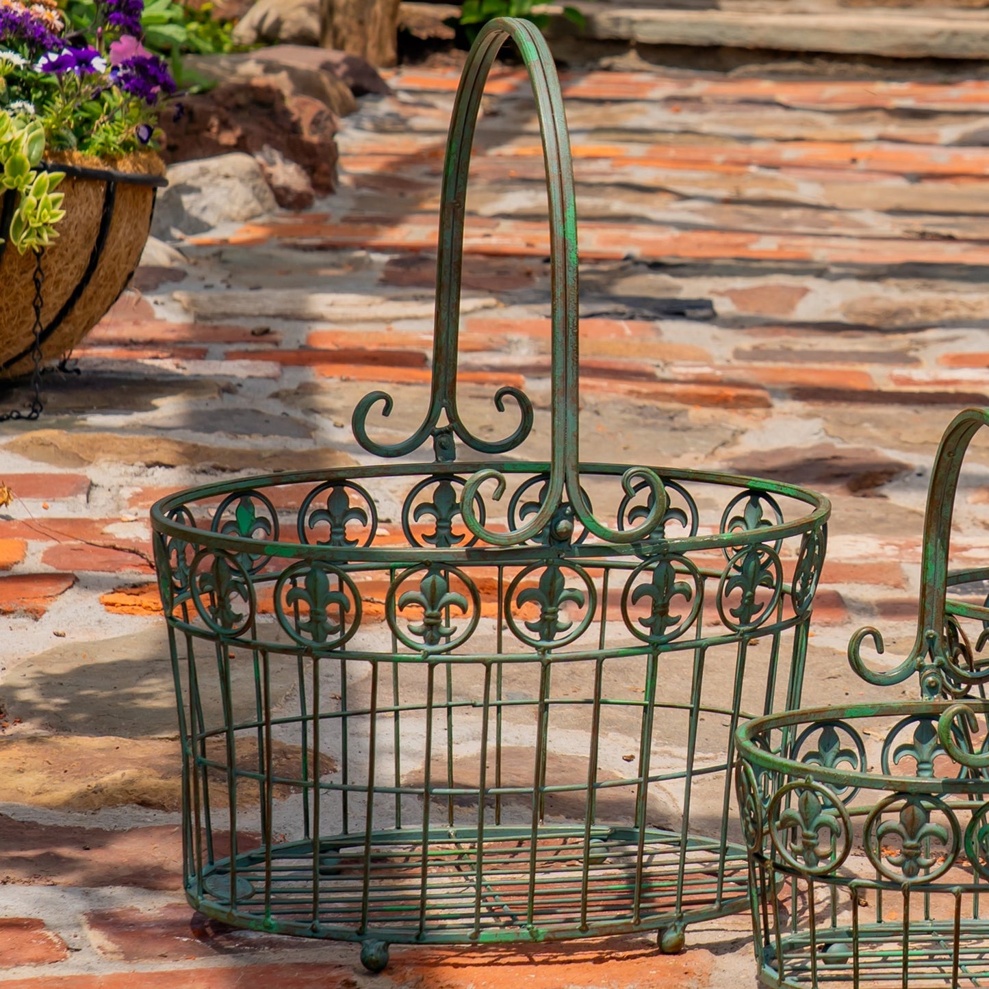 A green metal wire basket with an ornate scrollwork pattern and a tall arched handle, shown outdoors on a stone patio with a smaller matching basket partially visible at the right edge.