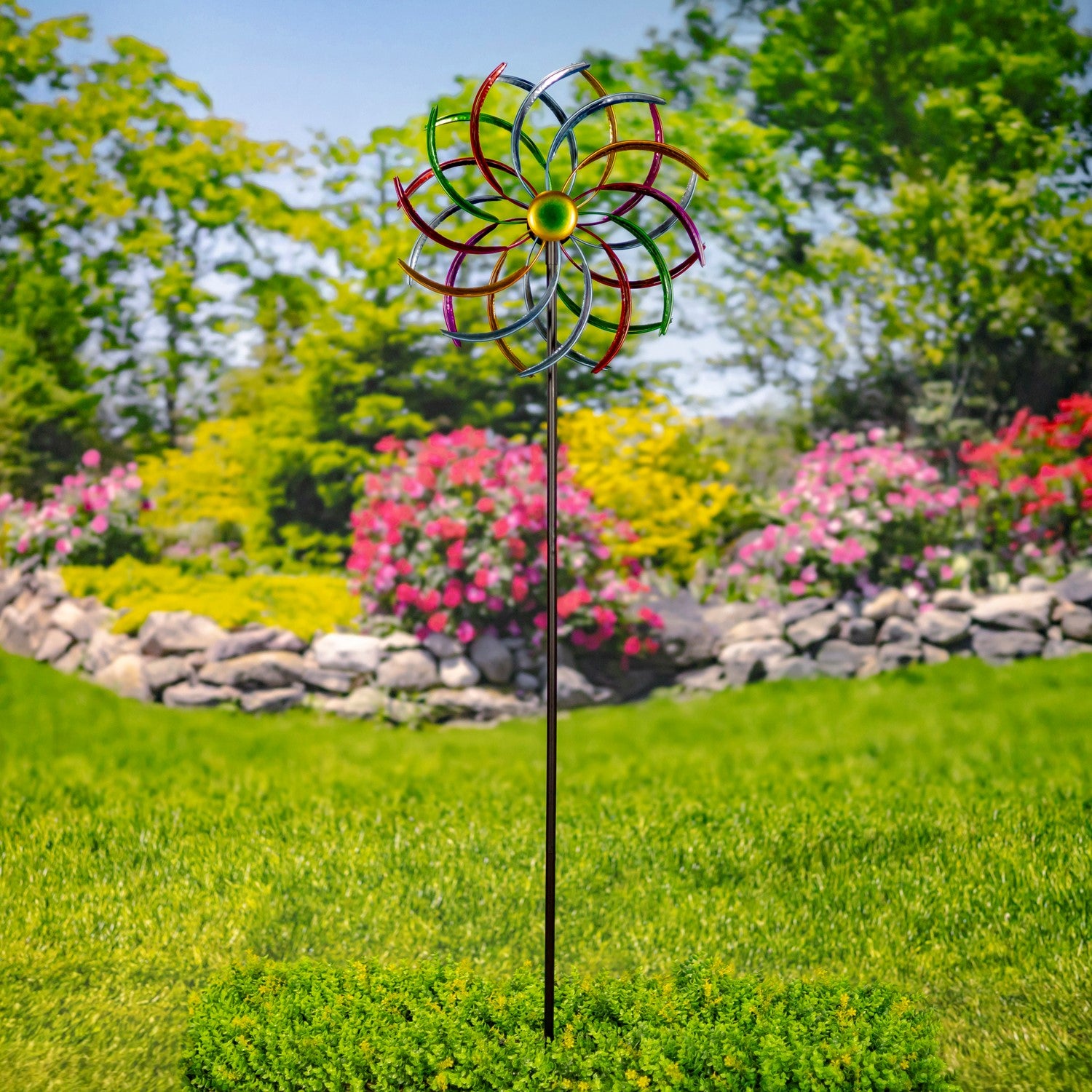 A metal garden wind spinner with a colorful spiral blade design and a yellow center, mounted on a black stake in a green grassy lawn with a blurred garden background.